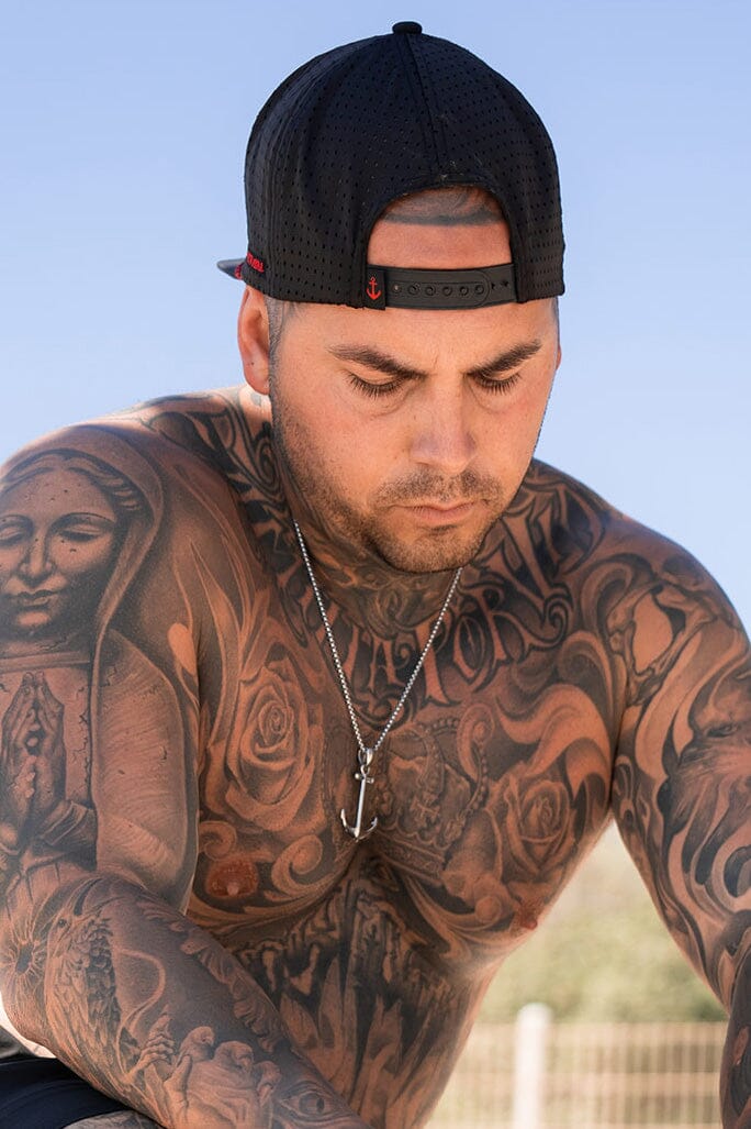 A man with extensive chest and arm tattoos gazes downwards, sporting a black baseball cap worn backwards and a Relentless Betrayal Anchor Drop Necklace, handcrafted with a 26-inch chain. The sky is clear blue in the background.