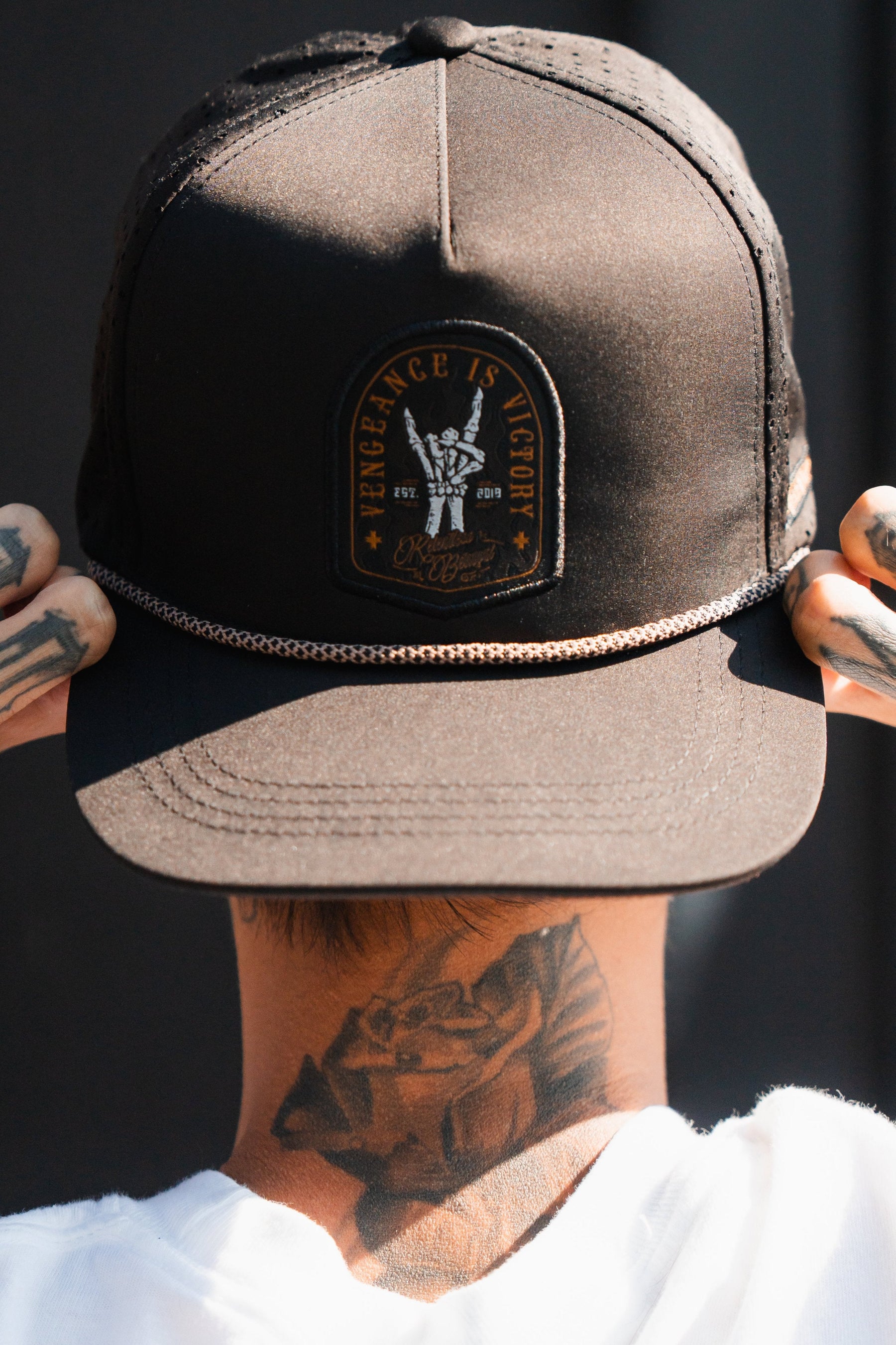 Seen from behind, exuding resilience, a person adjusts a Vengeance Snapback by Relentless Betrayal. Tattoos snake across their hands and neck, telling stories of strength as they grip the brim with determination.