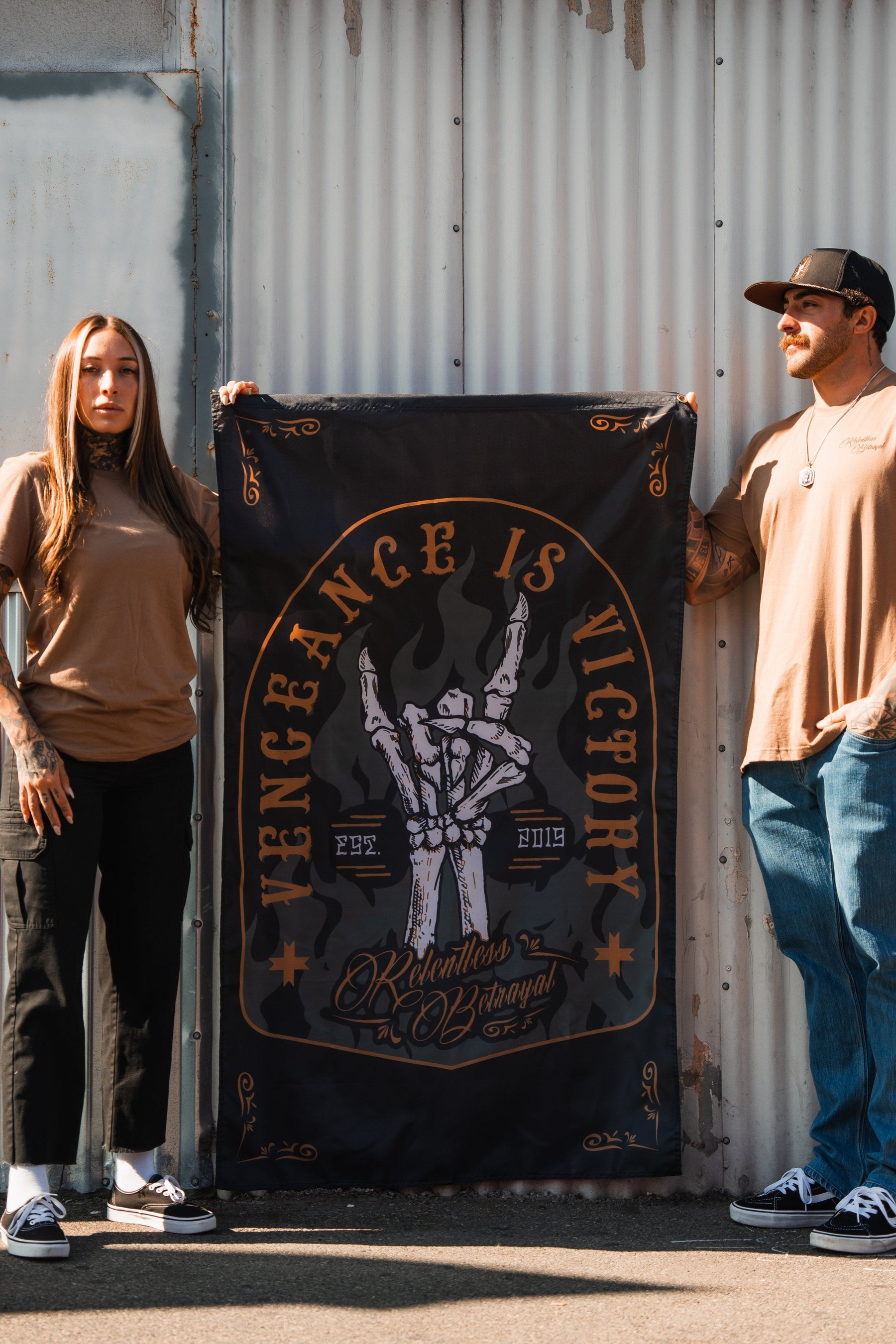 Two people hold a Vengeance Flag by Relentless Betrayal, featuring Vengeance is Victory with a skeletal hand illustration. Casually dressed, they confidently pose against a corrugated metal wall, symbolizing resilience and determination.