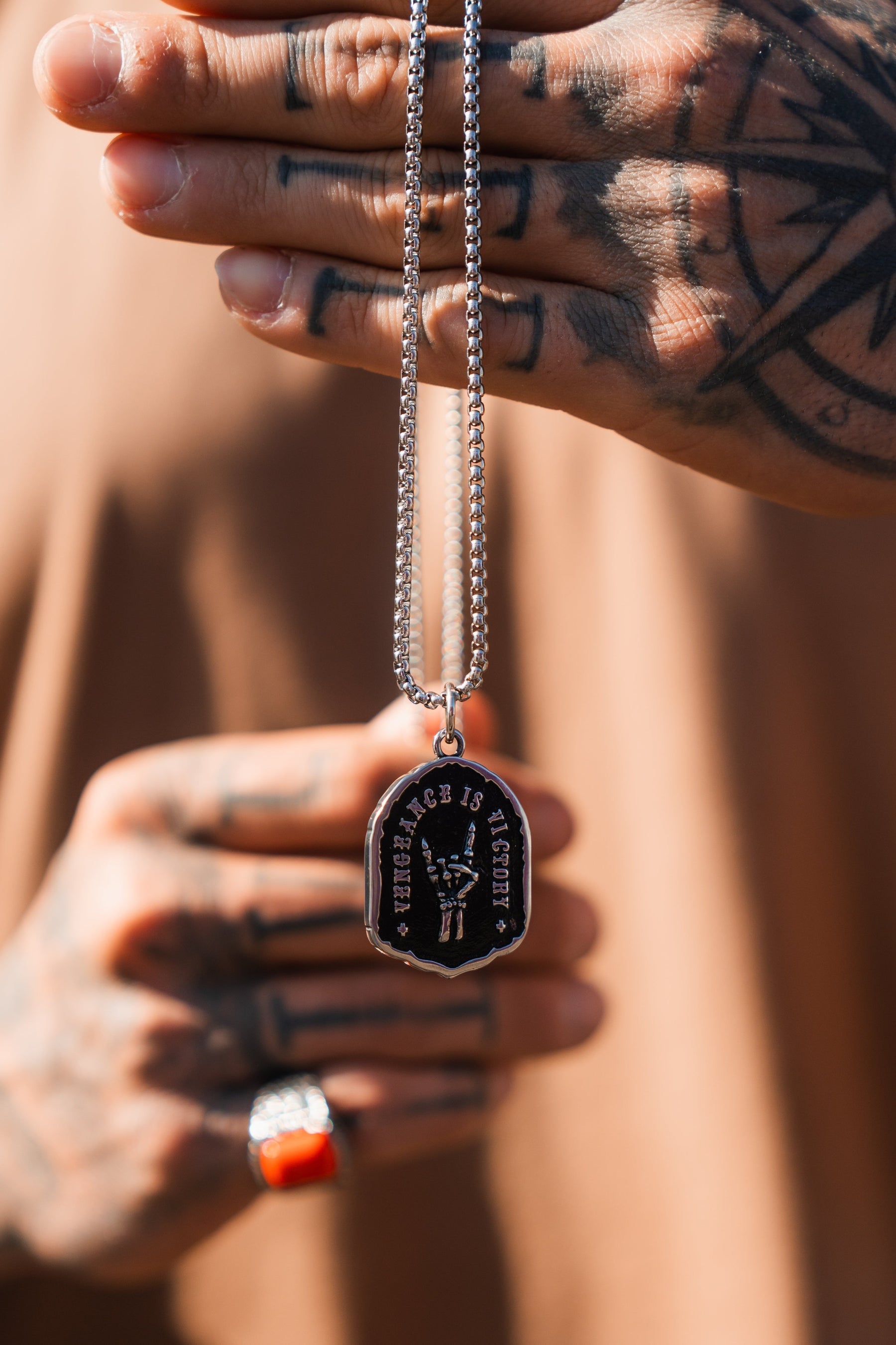 A tattooed hand holds the Vengeance Necklace by Relentless Betrayal, featuring a pendant that reads “Suffering is Inevitable,” symbolizing resilience. The focus is on the jewelry, while the person wears a beige top and a ring with a striking red stone.