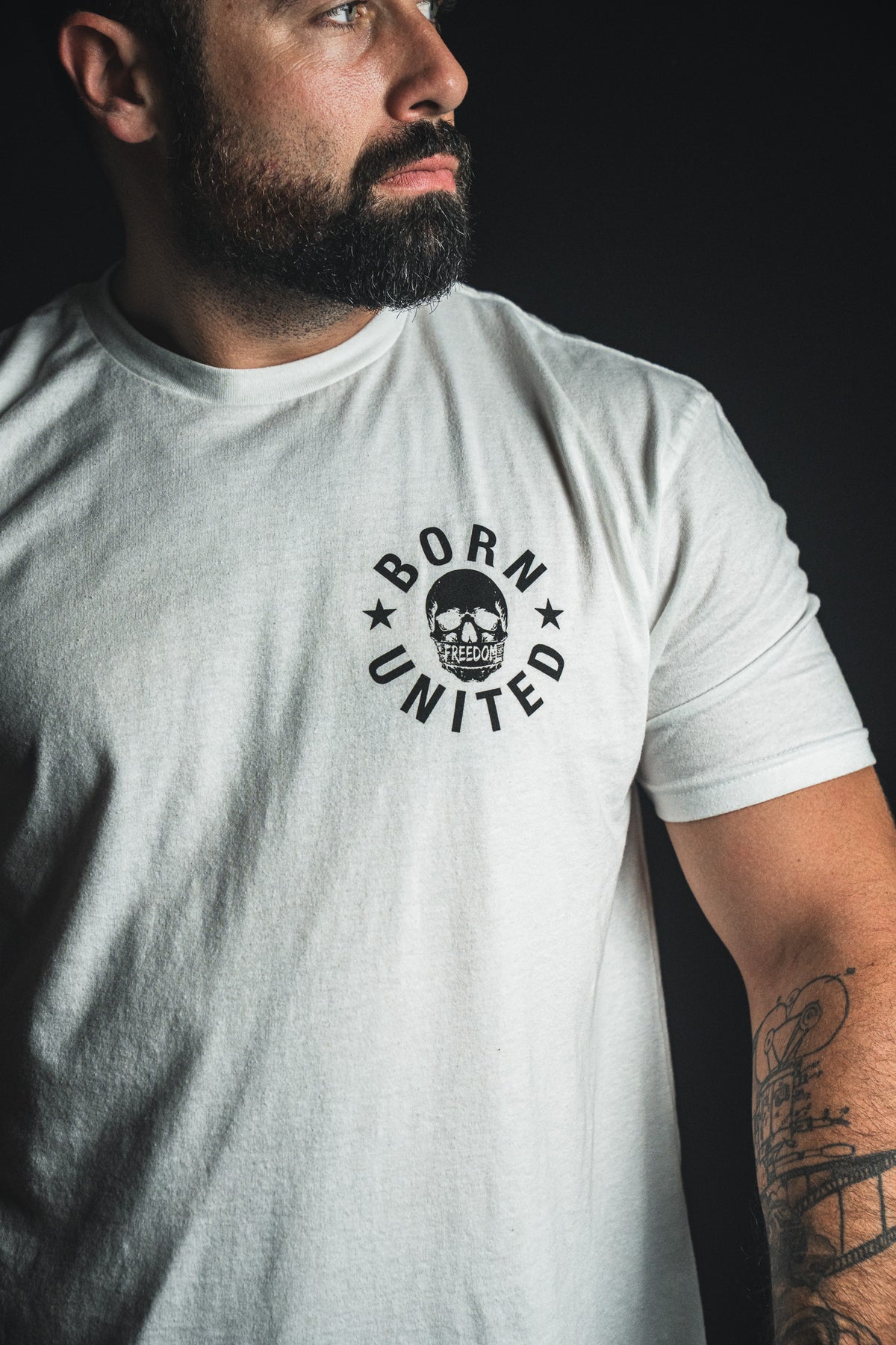 A bearded man wearing a “Freedom of Speech - White” t-shirt by Born United Private Label, featuring the signature BORN UNITED circular logo on the chest, stands against a dark background—a look that subtly reflects his commitment to Christian faith.