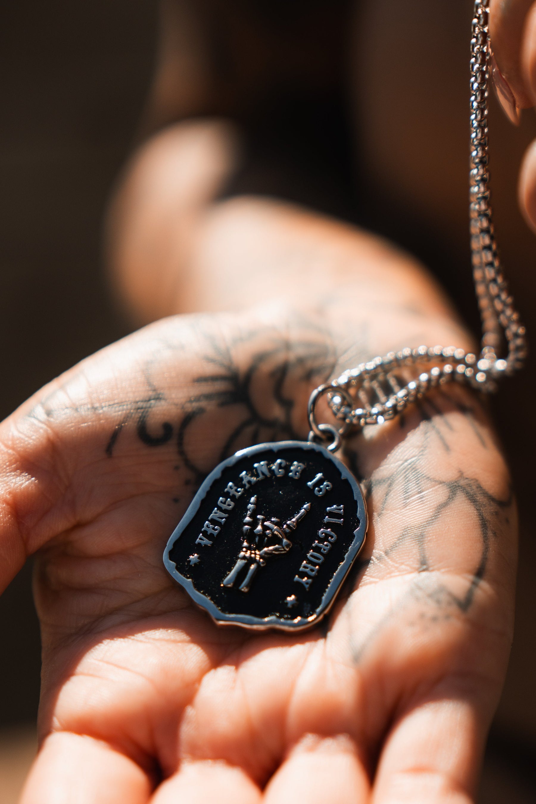 A tattooed hand, symbolizing resilience, holds the Vengeance Necklace by Relentless Betrayal. The pendant reads The King is Dead under a crown, casting empowering reflections as sunlight dances upon it.