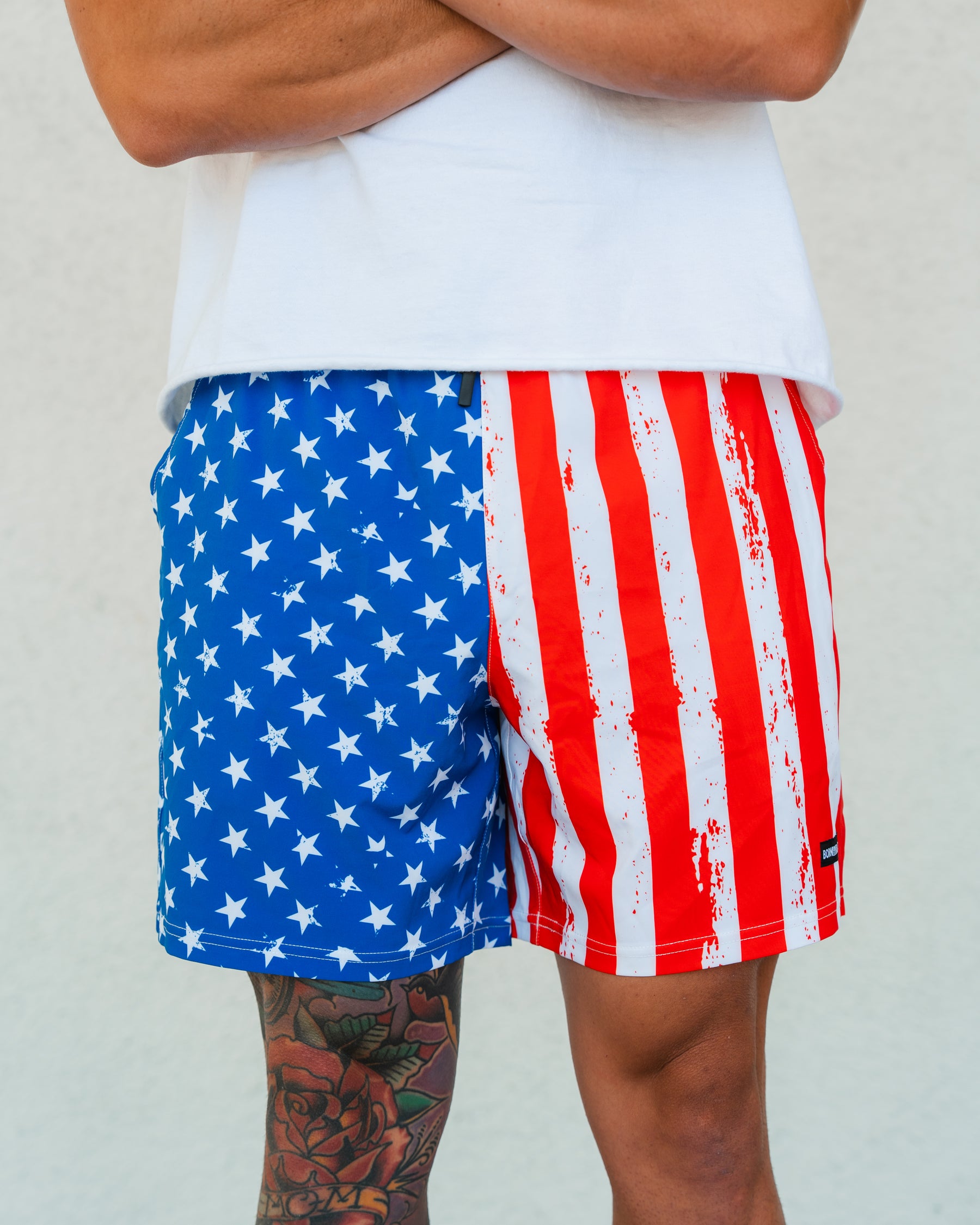 A person sports the Born United Private Label Mens Performance Hybrid Shorts 5.5 | Stars & Stripes, featuring an American flag pattern, paired with a plain white shirt; a flower tattoo is visible on their leg as they stand with crossed arms.