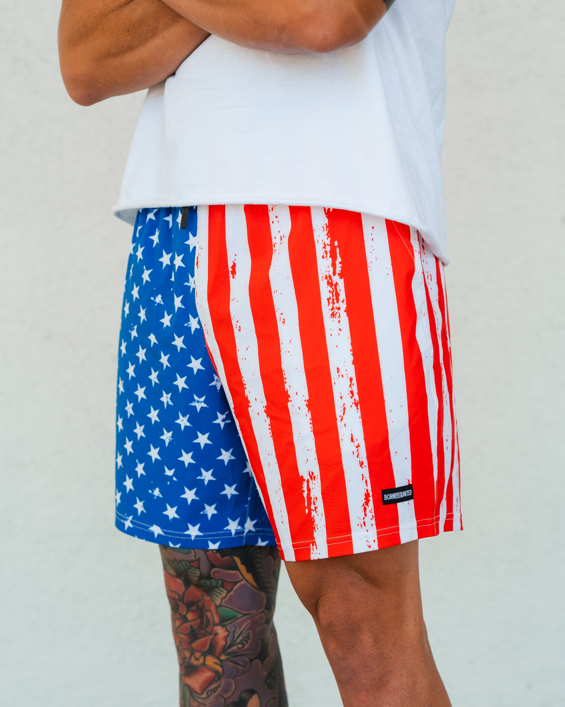 A person with crossed arms wears Born United Private Label Mens Performance Hybrid Shorts 5.5 | Stars & Stripes, featuring a quick-drying American flag design, paired with a white T-shirt. A colorful tattoo is visible on their lower leg.