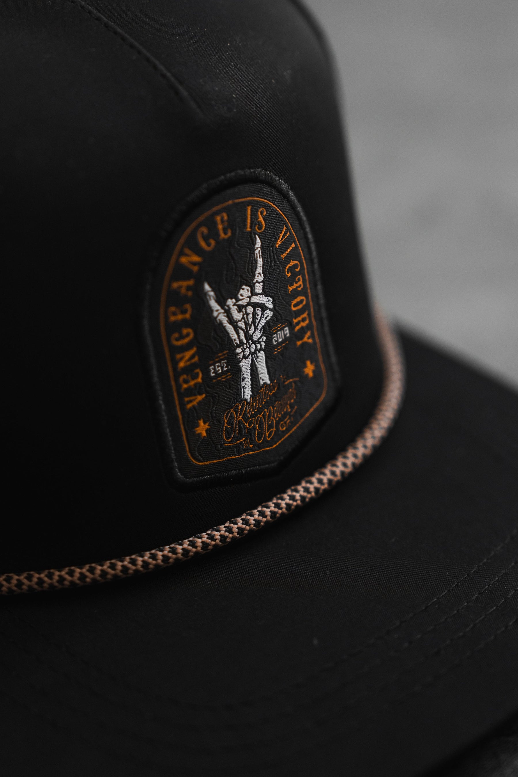 The Vengeance Snapback by Relentless Betrayal showcases an empowering black cap with an arch-shaped patch reading VENGEANCE IS VICTORY, featuring a hand with a trident. It includes SFC and dates 2017-2019, complemented by a braided cord on the brim, symbolizing resilience.