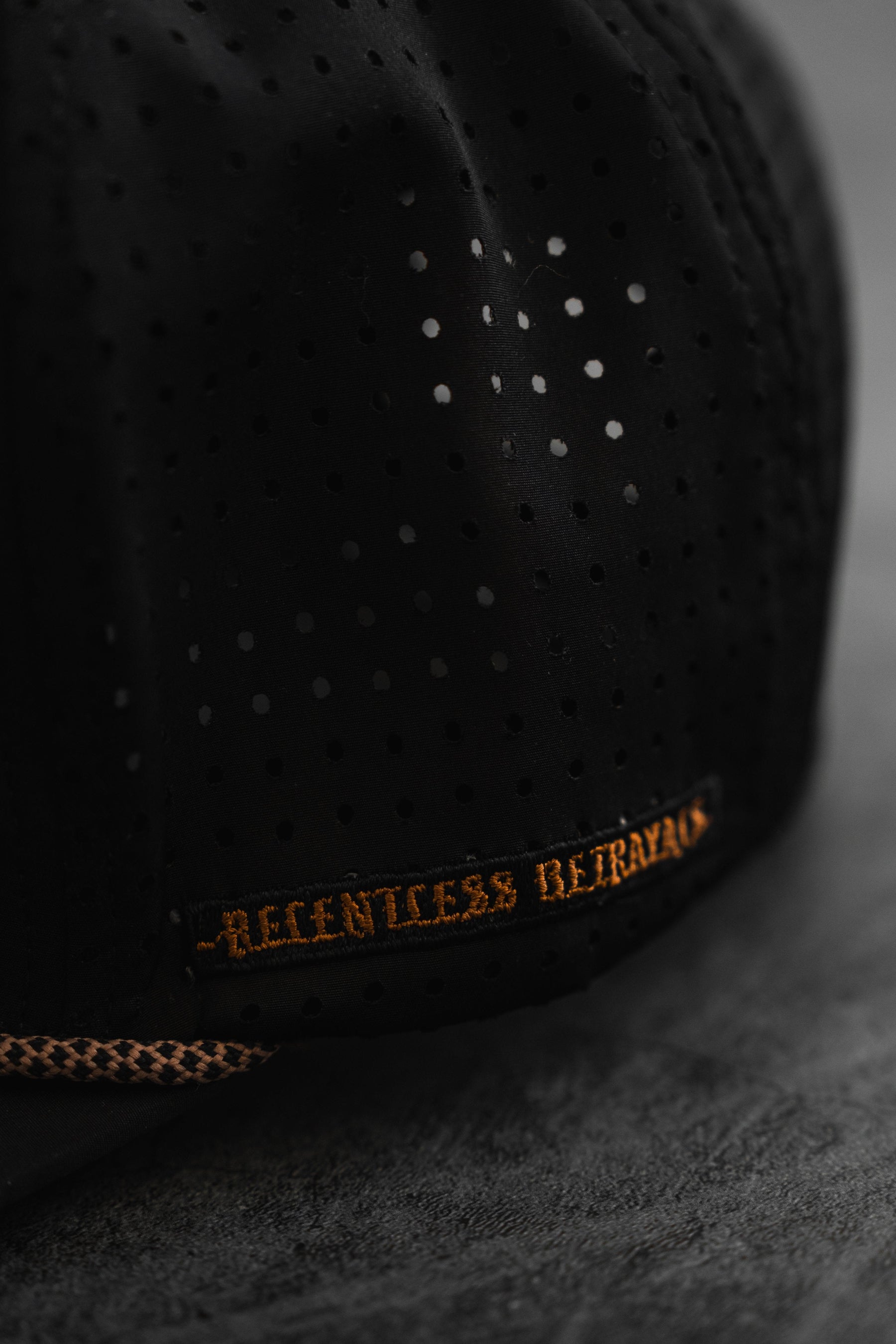 Close-up of the Vengeance Snapback by Relentless Betrayal, featuring a black perforated fabric with subtle polka dots and an embroidered brown RELENTLESS BETRAYAL phrase along the edge, set against a textured gray surface.