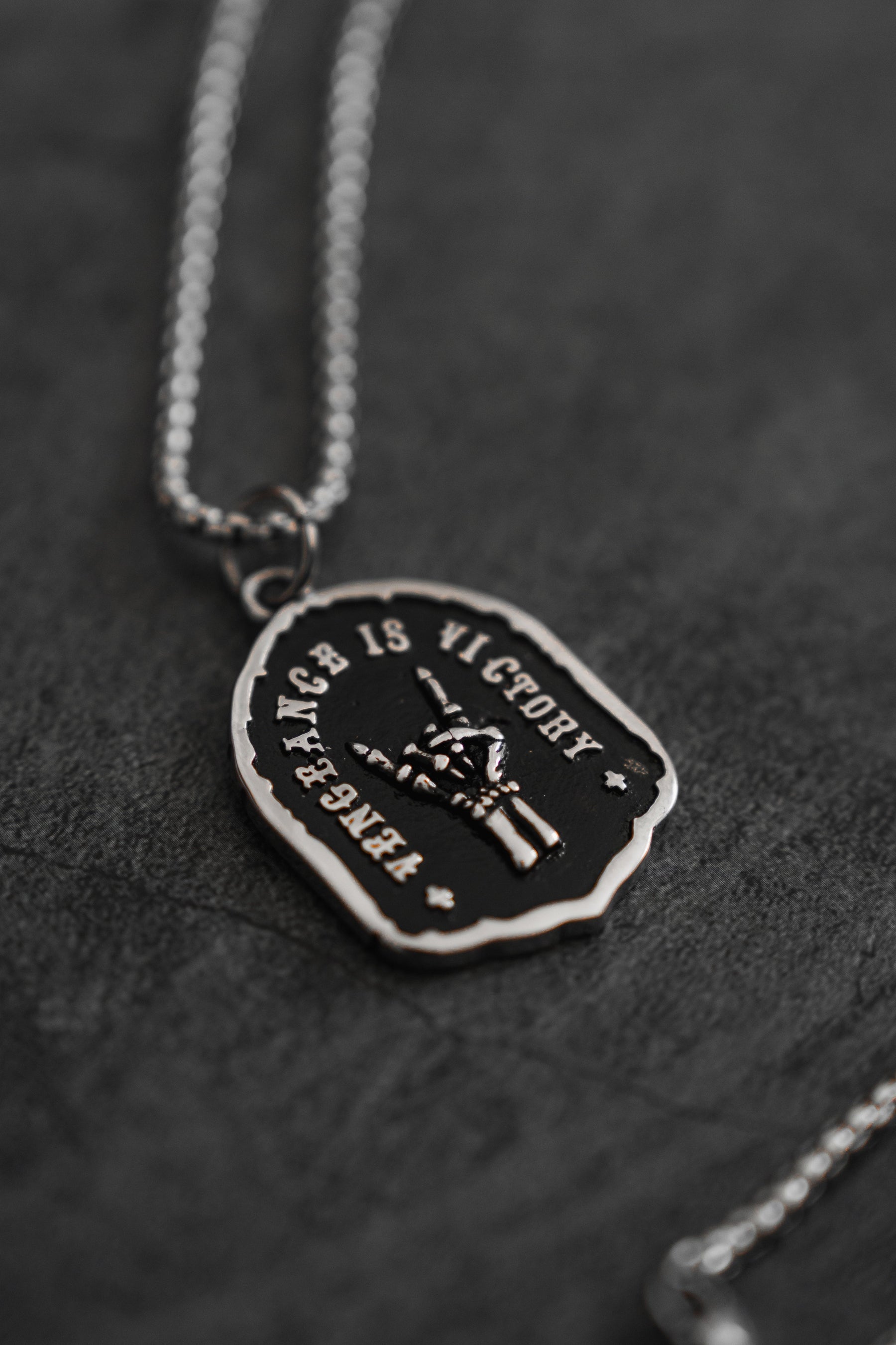 The Vengeance Necklace by Relentless Betrayal features a silver peace sign hand pendant engraved with Vengeance is Victory on a silver chain, set against a dark, textured background.
