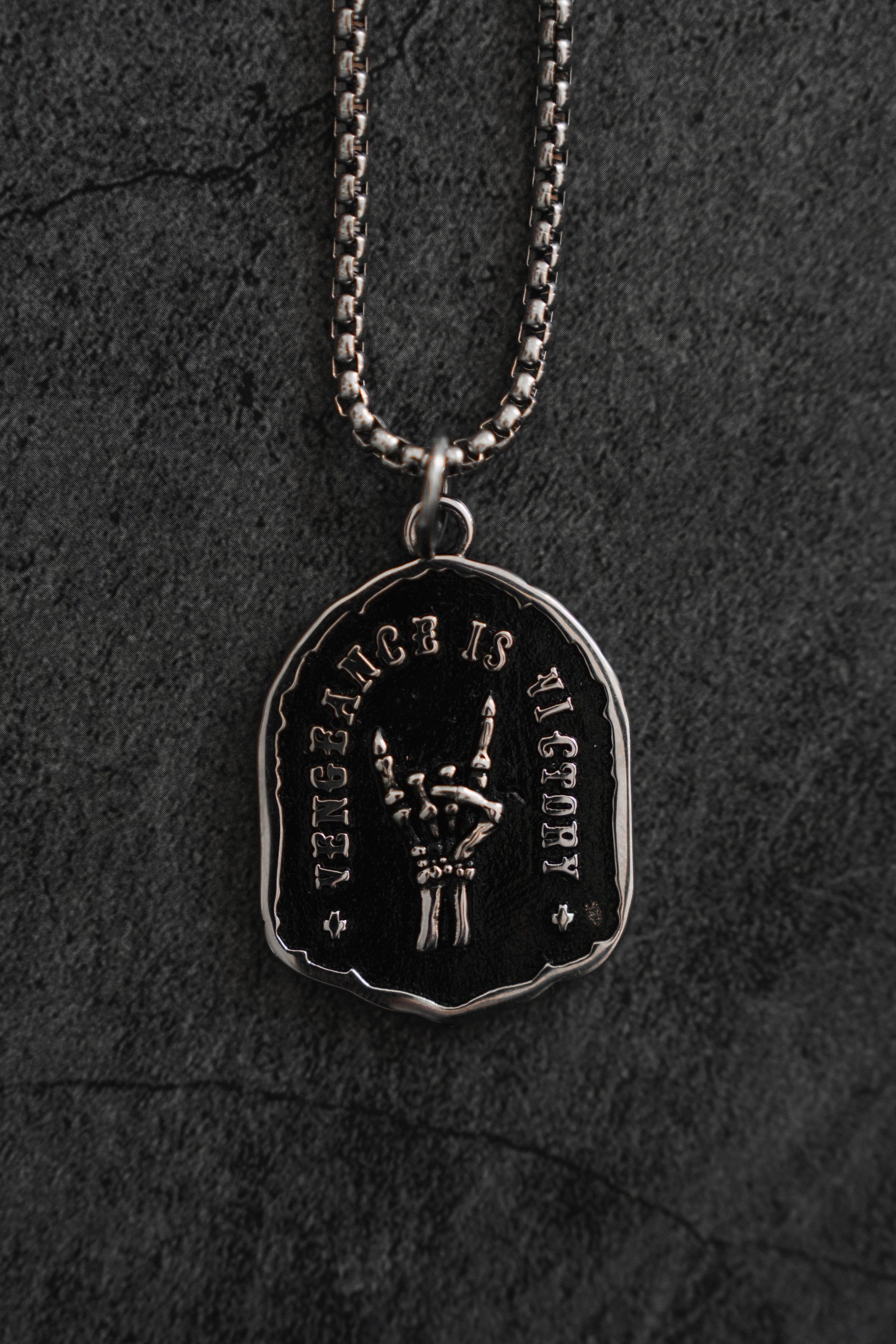 The Vengeance Necklace from Relentless Betrayal is displayed against a dark background, featuring a round, rustic silver pendant. The engraved skeletal arm holding a spear is encircled by VIGILANCE IS VICTORY, symbolizing resilience and empowerment.