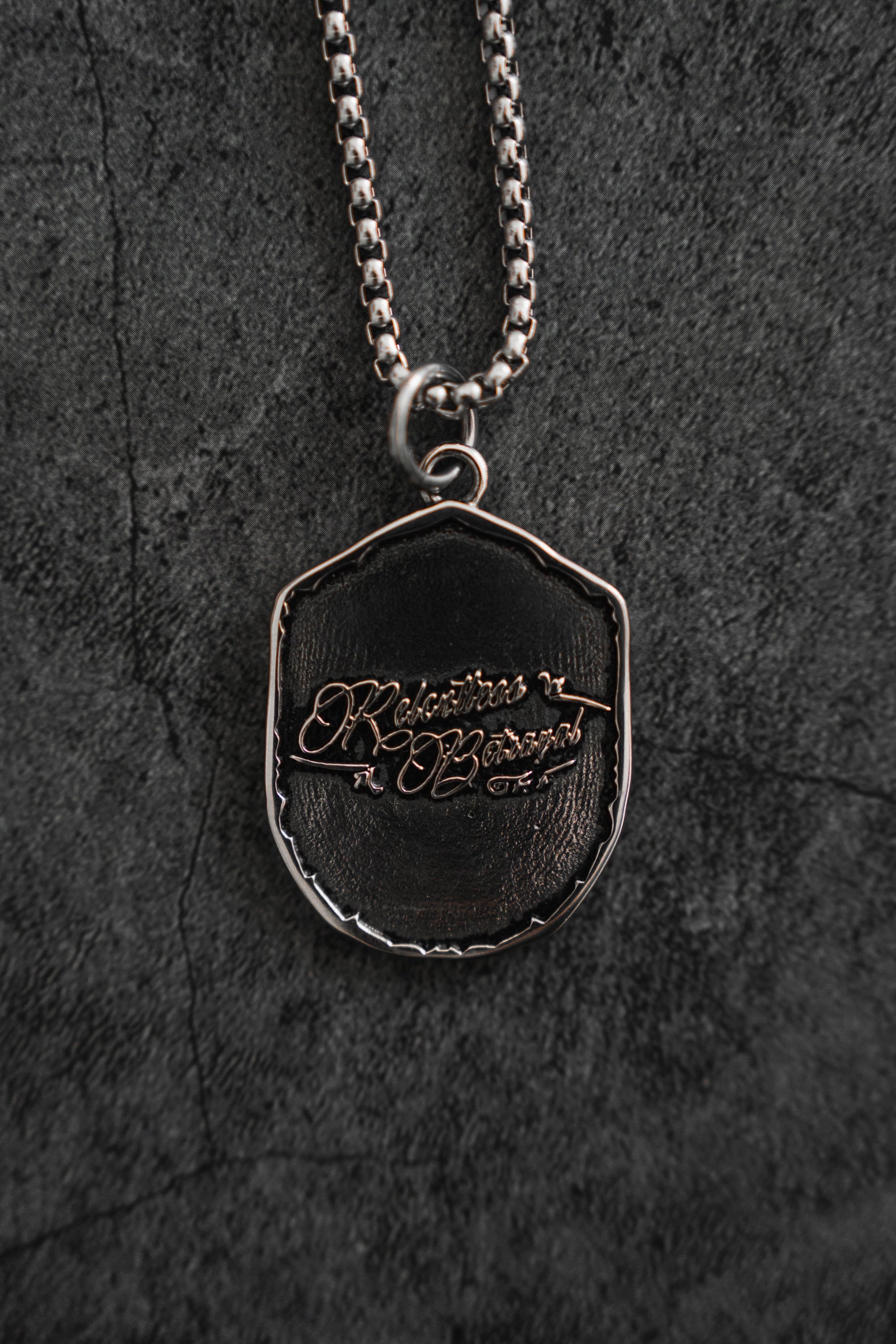 The Vengeance Necklace by Relentless Betrayal features a silver pendant engraved with Vengeance is Victory in a decorative script. Its intricate design, set against a textured dark background, symbolizes resilience and empowerment.