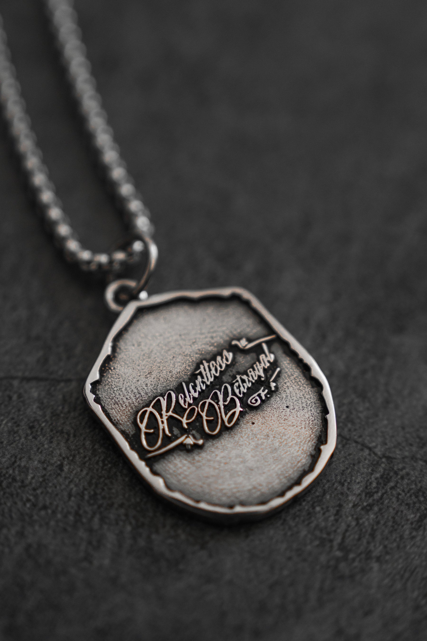The Vengeance Necklace by Relentless Betrayal features a textured silver chain with an oval pendant. Engraved with Restless & Obscure in a stylized font, it symbolizes resilience and empowerment through artistic elegance, all resting on a dark surface.