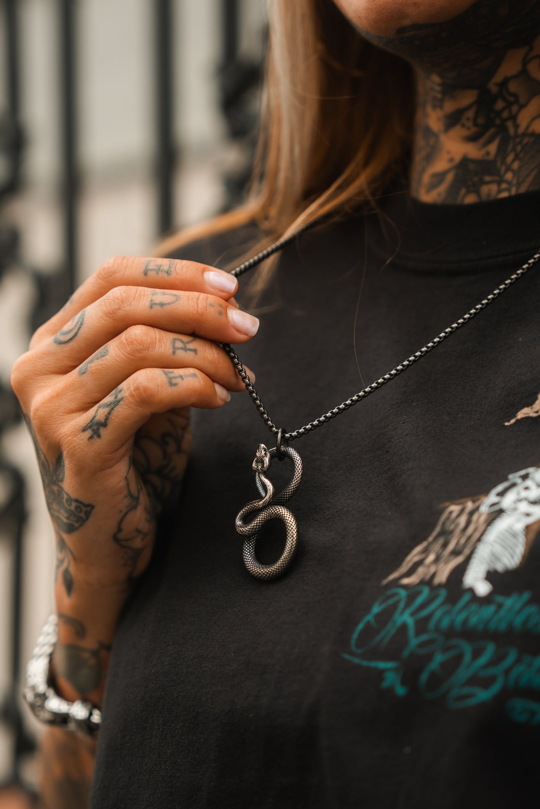 Clad in a black shirt with a graphic design, an individual showcases the Slither Necklace by Relentless Betrayal. The serpentine pendant is complemented by tattoos on their hands and neck. A blurred railing in the background adds to the accessorys enigmatic allure.