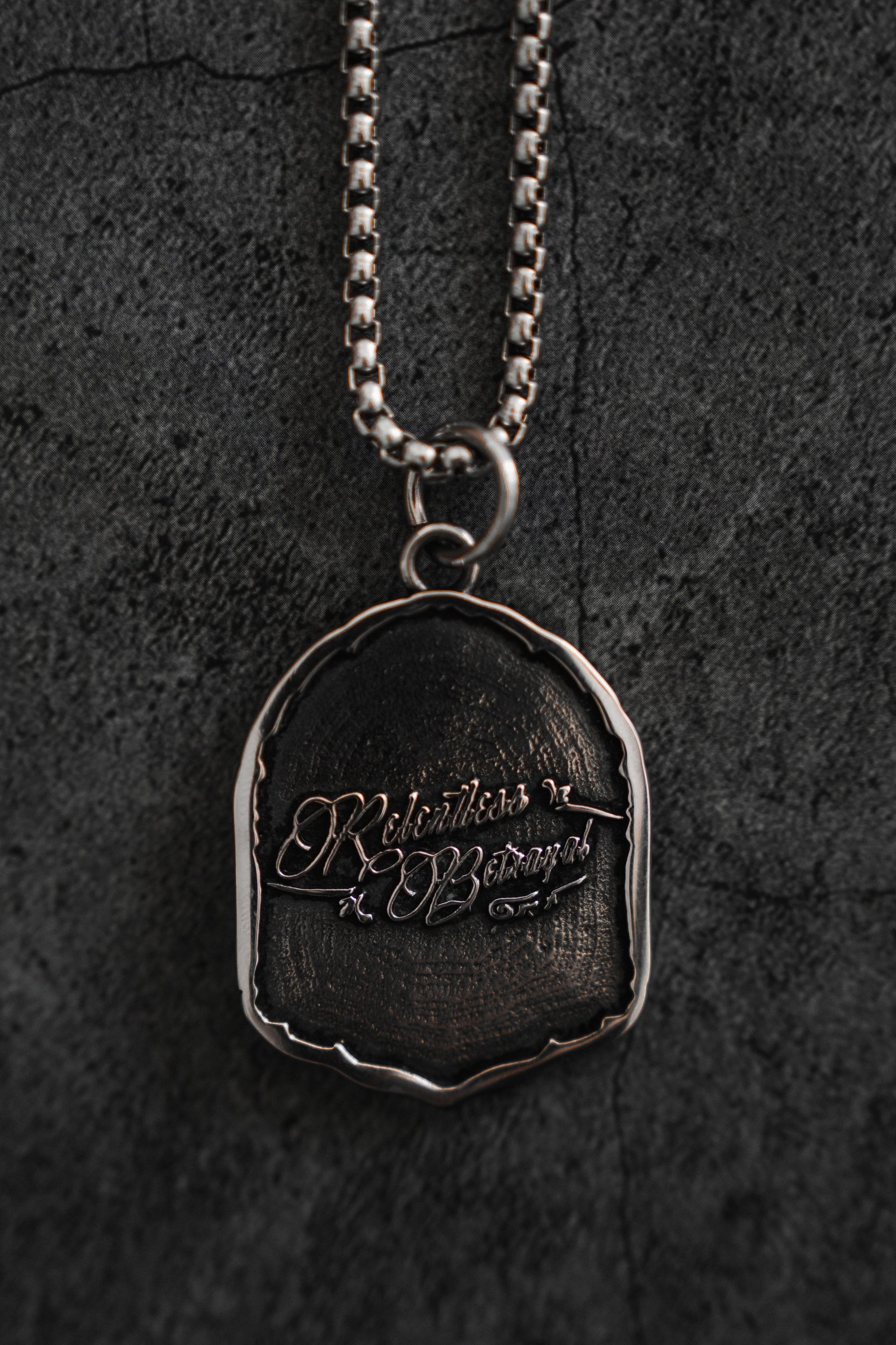 The Vengeance Necklace by Relentless Betrayal features an oval silver pendant with intricate script on a dark, textured surface symbolizing resilience, gracefully hanging from a chain against a dark, mottled background.