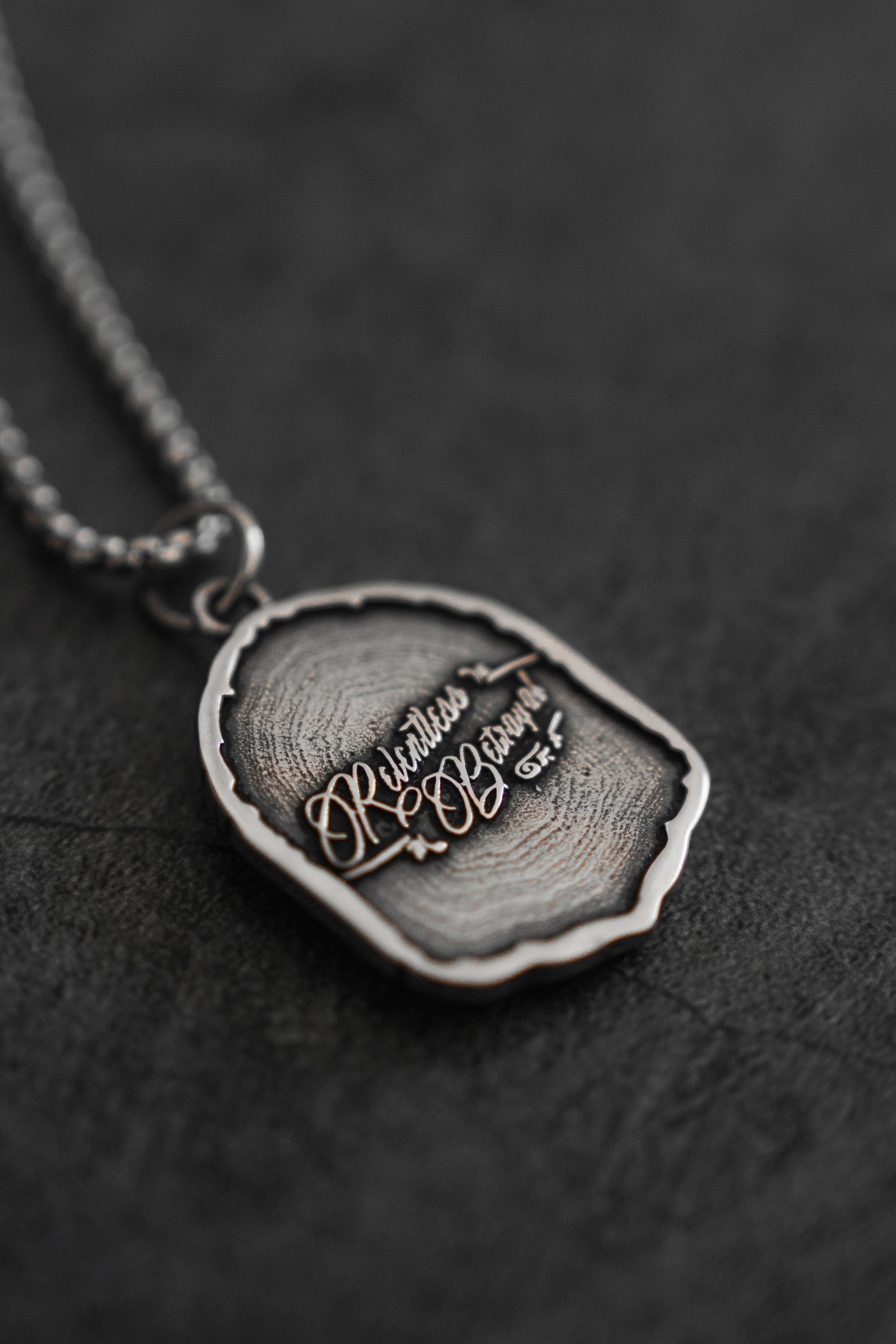The Vengeance Necklace by Relentless Betrayal features a silver pendant with an irregular, textured design. Engraved with Relentless, Be Brave, it symbolizes resilience and empowerment, set against a dark, textured background.