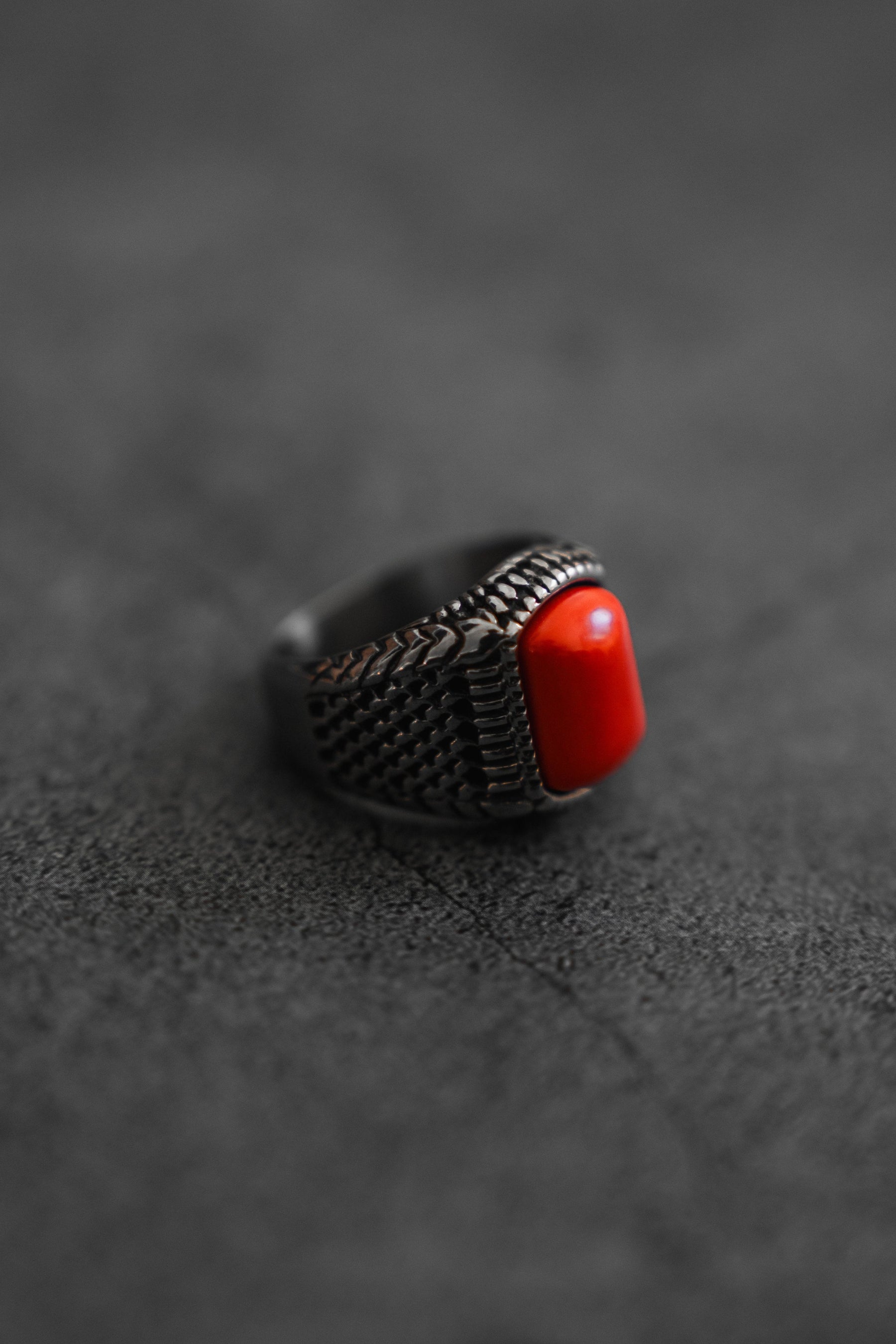 The Relentless Betrayal Hell Fire Ring, a handcrafted silver piece with intricate rope detailing and a large rectangular red stone, is elegantly perched on a dark, textured surface.