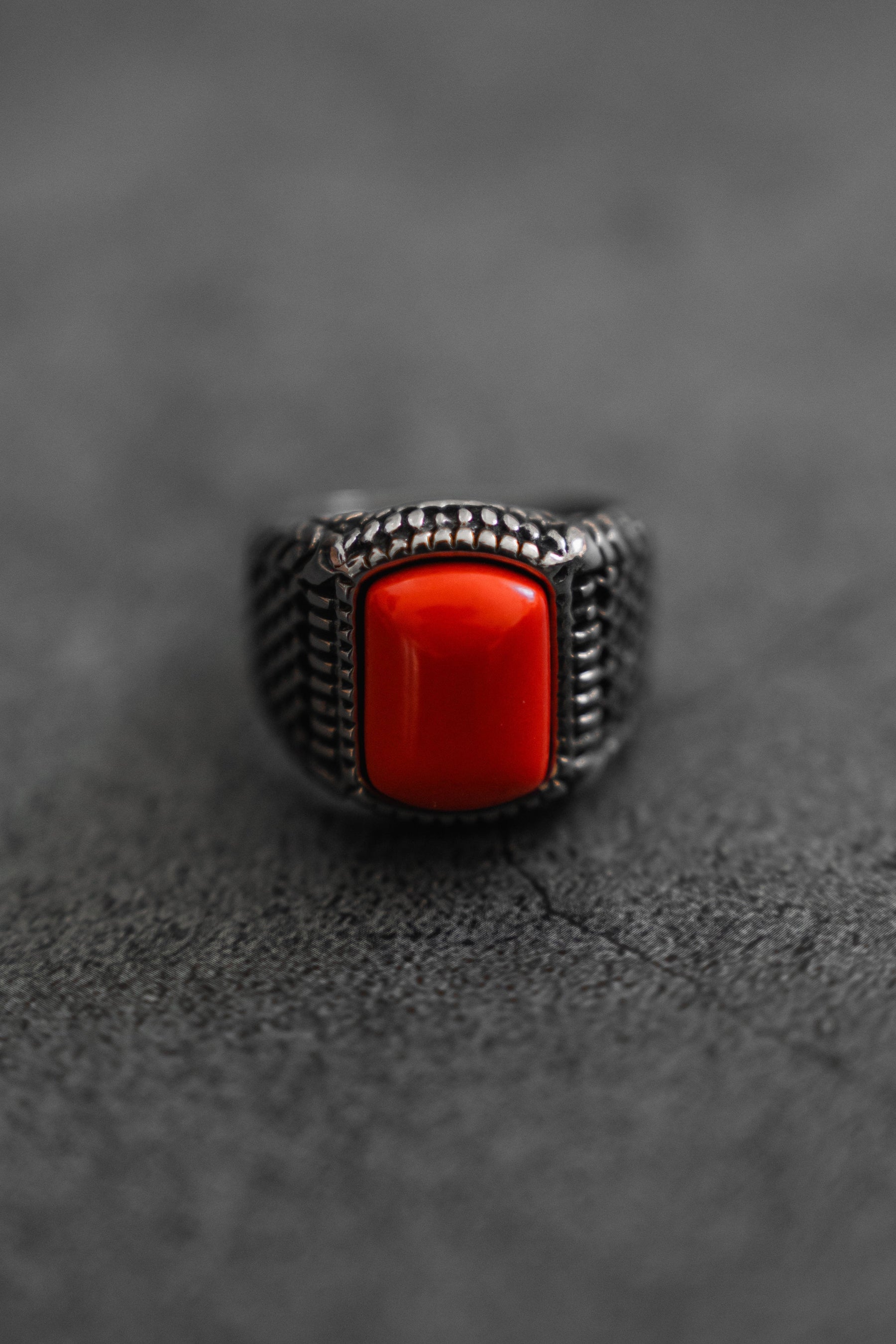 The handcrafted Hell Fire Ring by Relentless Betrayal features intricate detailing with a prominent rectangular red stone, resting elegantly on a textured gray surface and exuding timeless allure.
