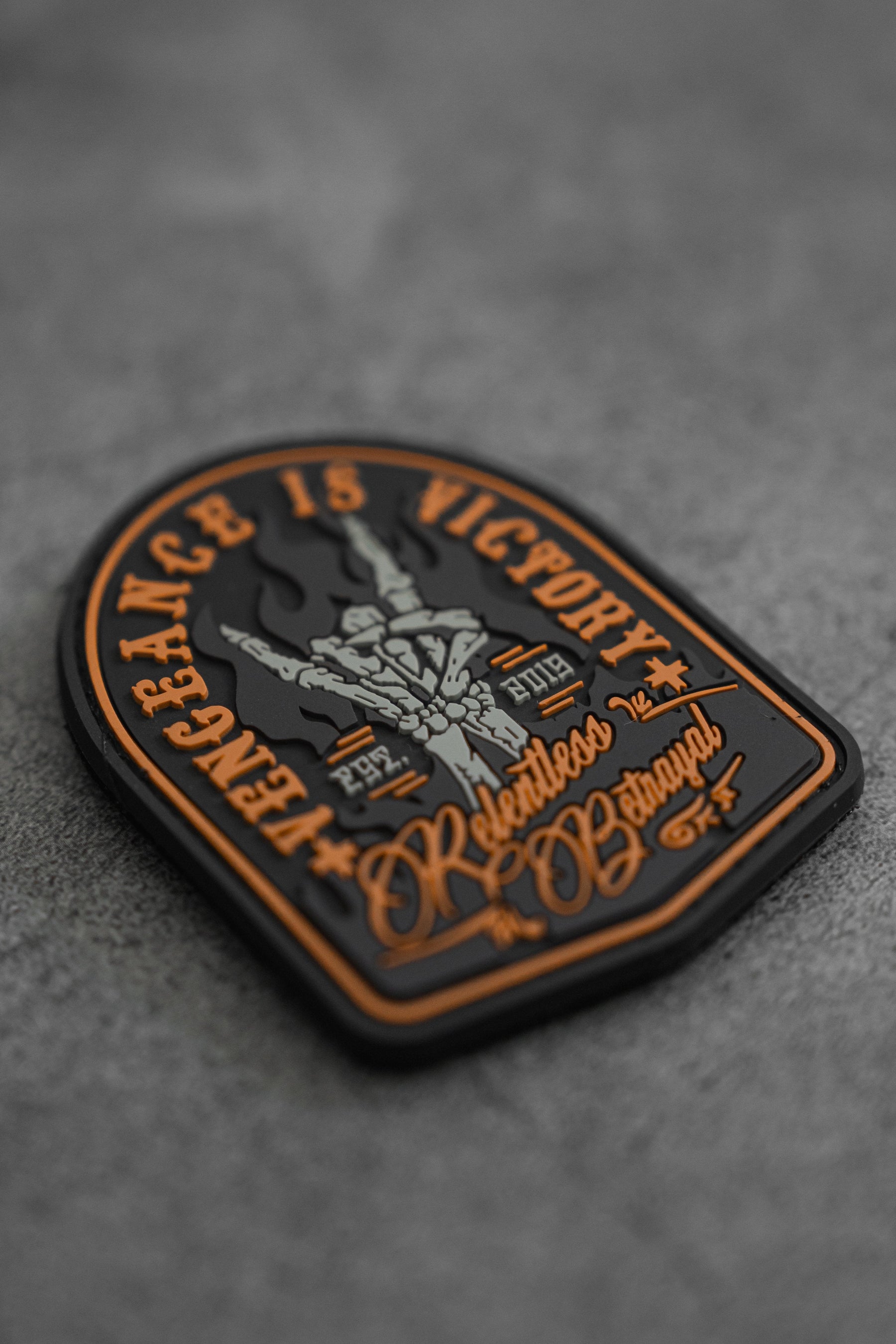 The Vengeance Patch by Relentless Betrayal features a black and orange design on gray, depicting a skeletal hand with two fingers raised. It includes the text Vengeance is Victory and Relentless Betrayal, conveying resilience with smaller inscriptions below.