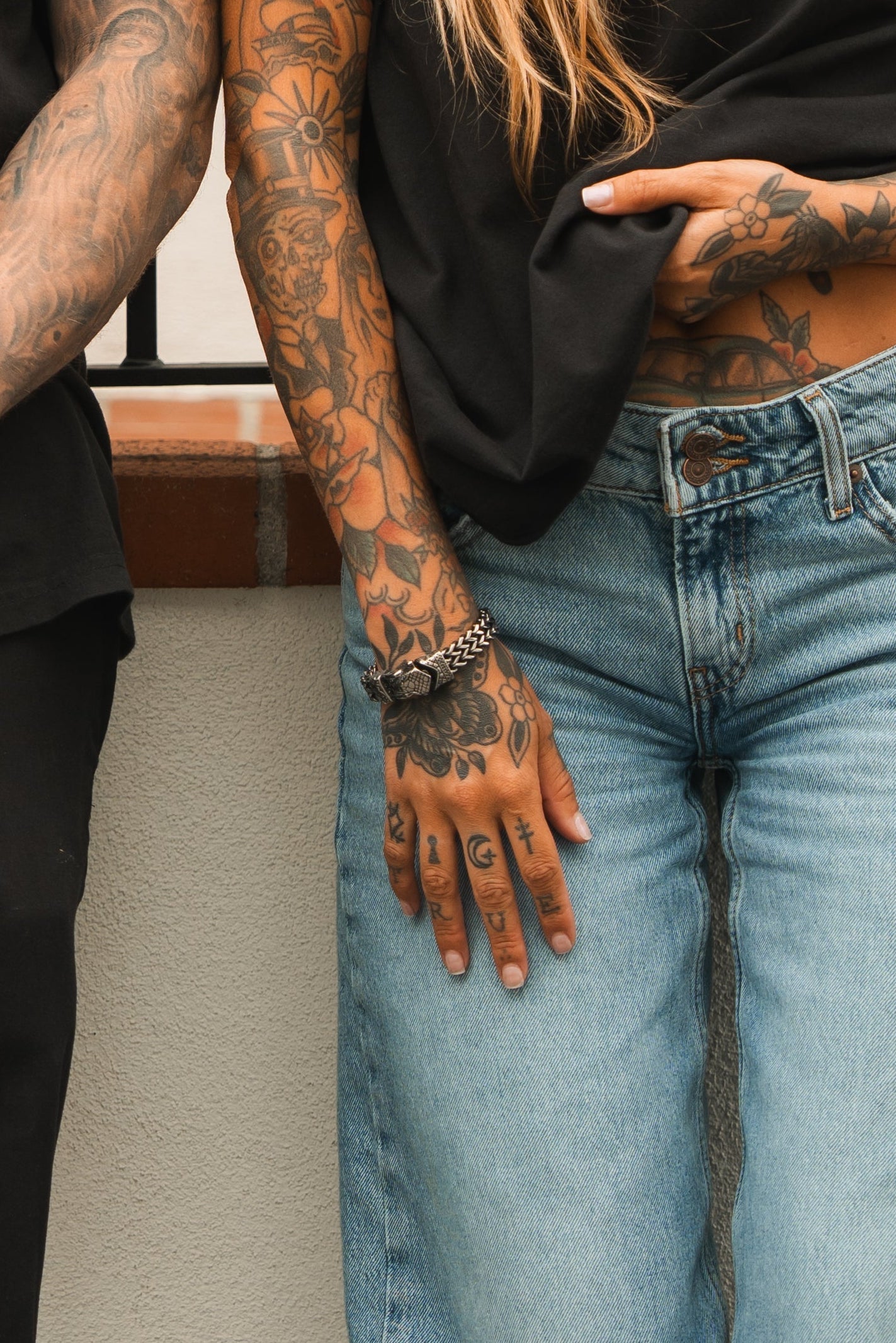 A person with tattooed arms and hands is in light blue jeans and a black shirt. Their left hand, adorned with tattoos, rests on their leg, showcasing the Relentless Betrayal Viper Bracelet. Another tattooed arm is partially visible to the left.