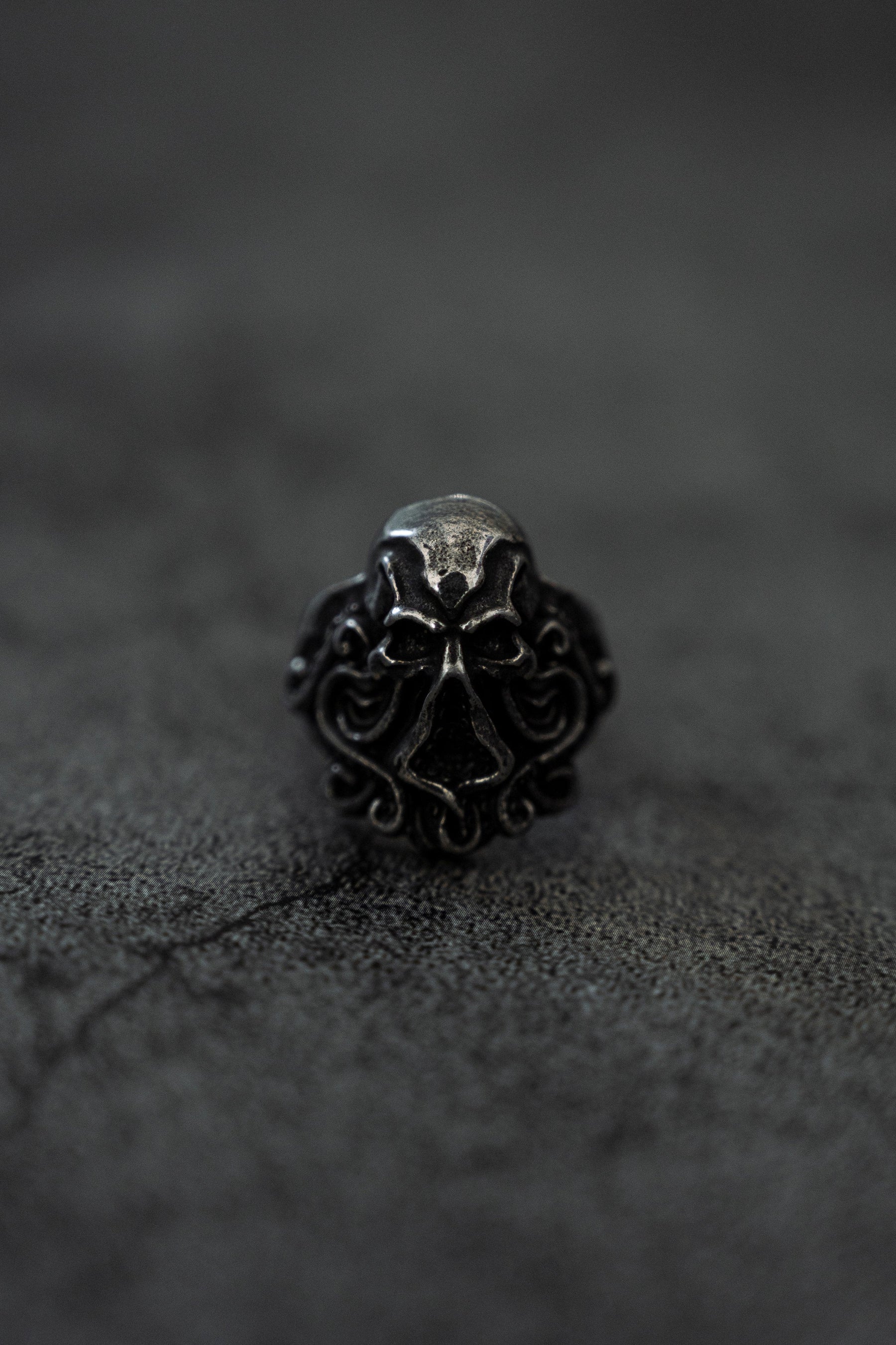 A close-up of The Kraken Ring by Relentless Betrayal, a stainless steel ring with an ornate skull design set against a dark, textured background. The skull features intricate patterns evoking kraken tentacles, creating a gothic aesthetic.