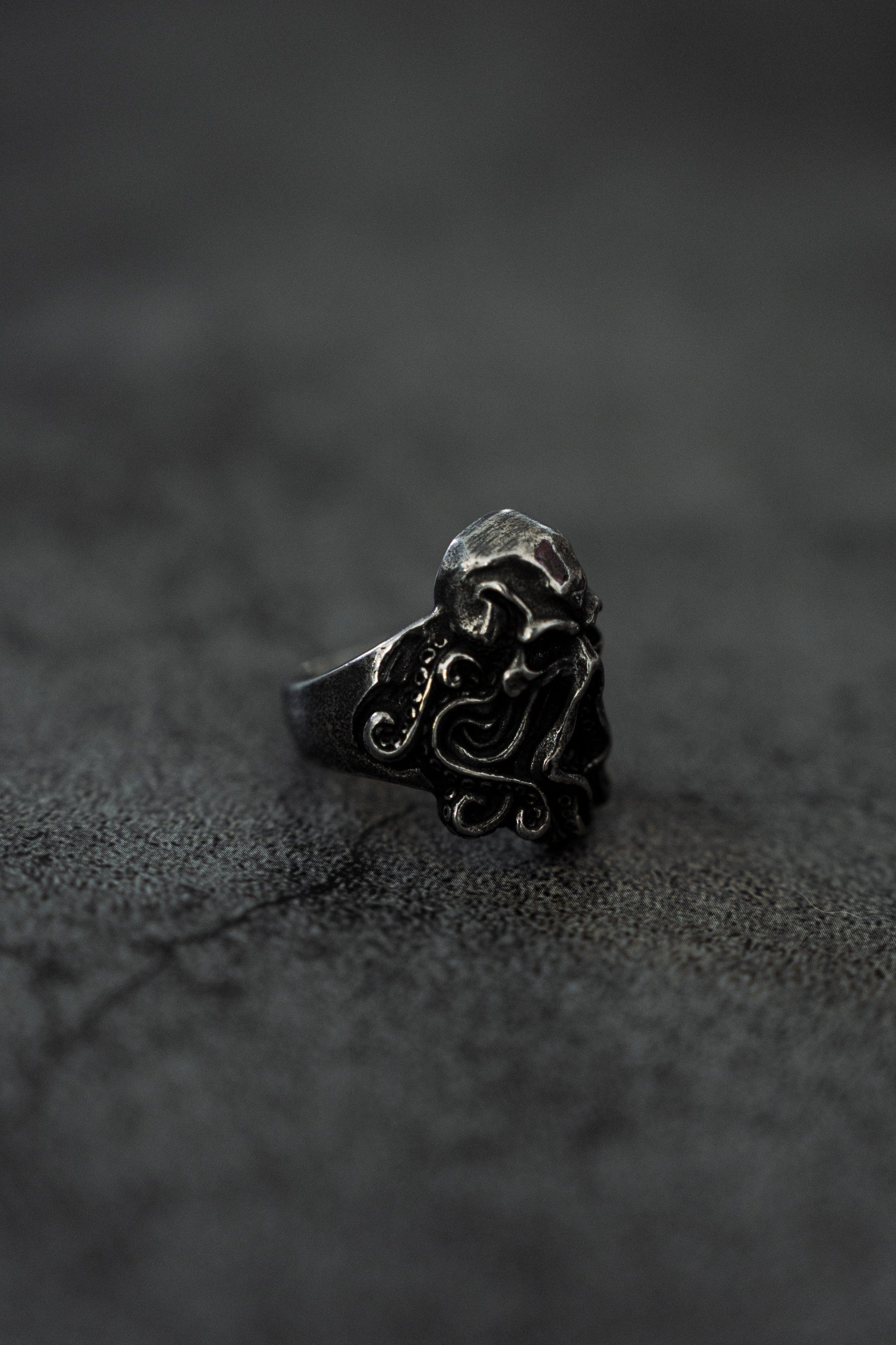 The Kraken Ring by Relentless Betrayal, a dark stainless steel ring with intricate swirling patterns inspired by Kraken tales, rests on a textured gray surface. Its handcrafted details stand out against the muted background, emphasizing its decorative elements.