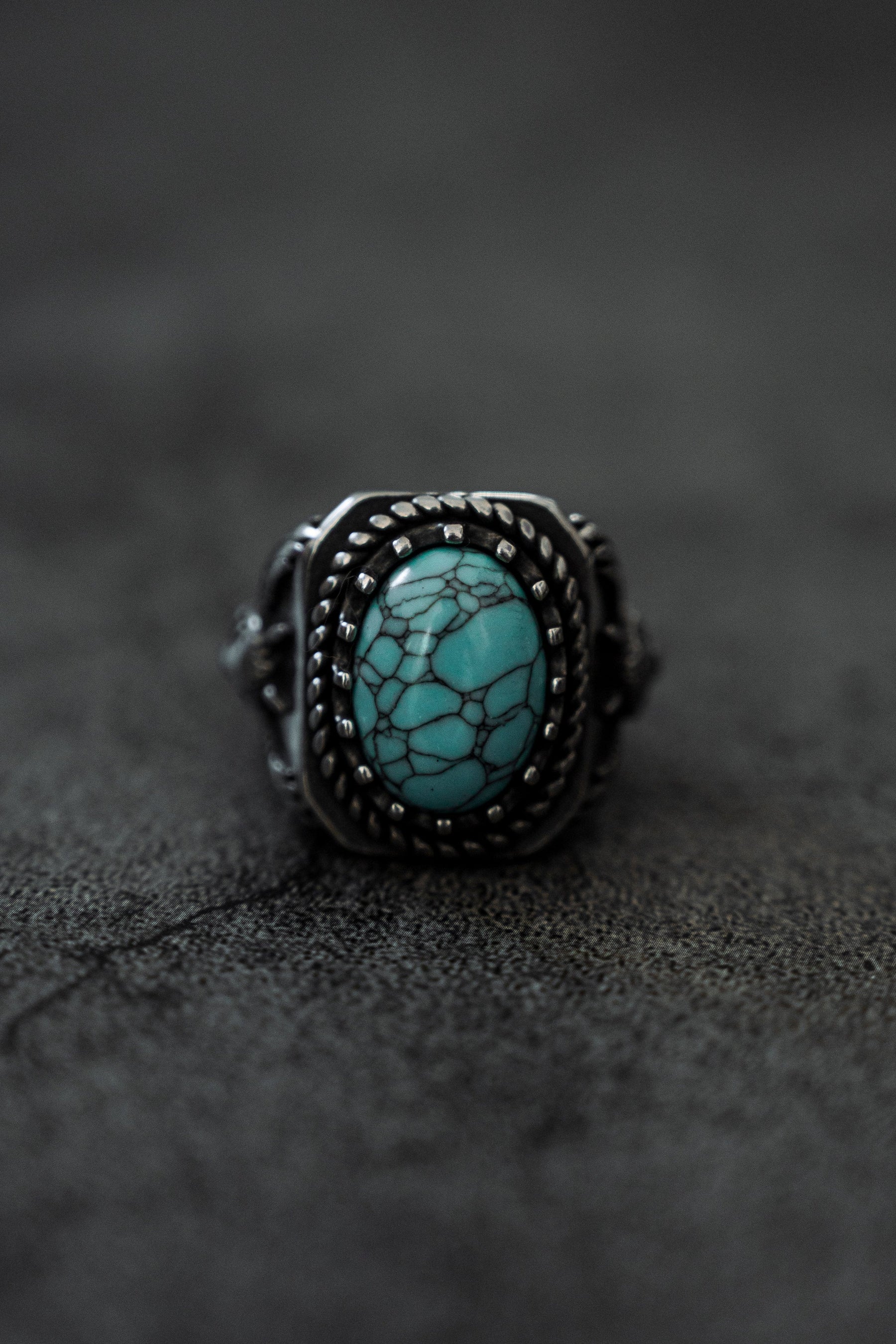 A vintage-inspired Relentless Betrayal Royal Guardian Ring showcasing a large oval turquoise stone with a web-like pattern, set in an intricately detailed band, rests on a dark textured surface.