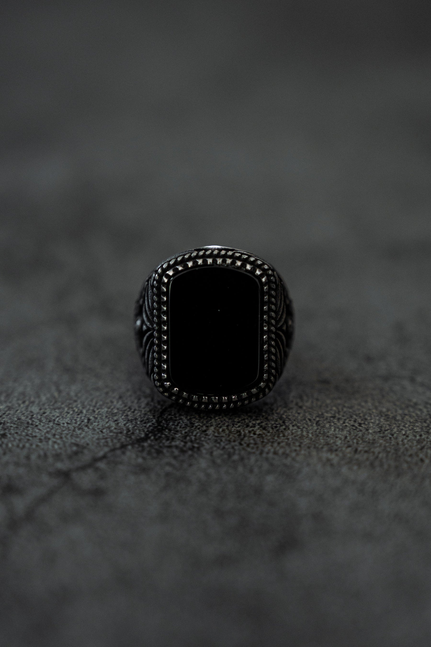 The Monarch Ring by Relentless Betrayal is a stainless steel piece featuring a rectangular black stone and detailed band, set against a textured dark surface casting a subtle shadow to embody transformation and elegance.