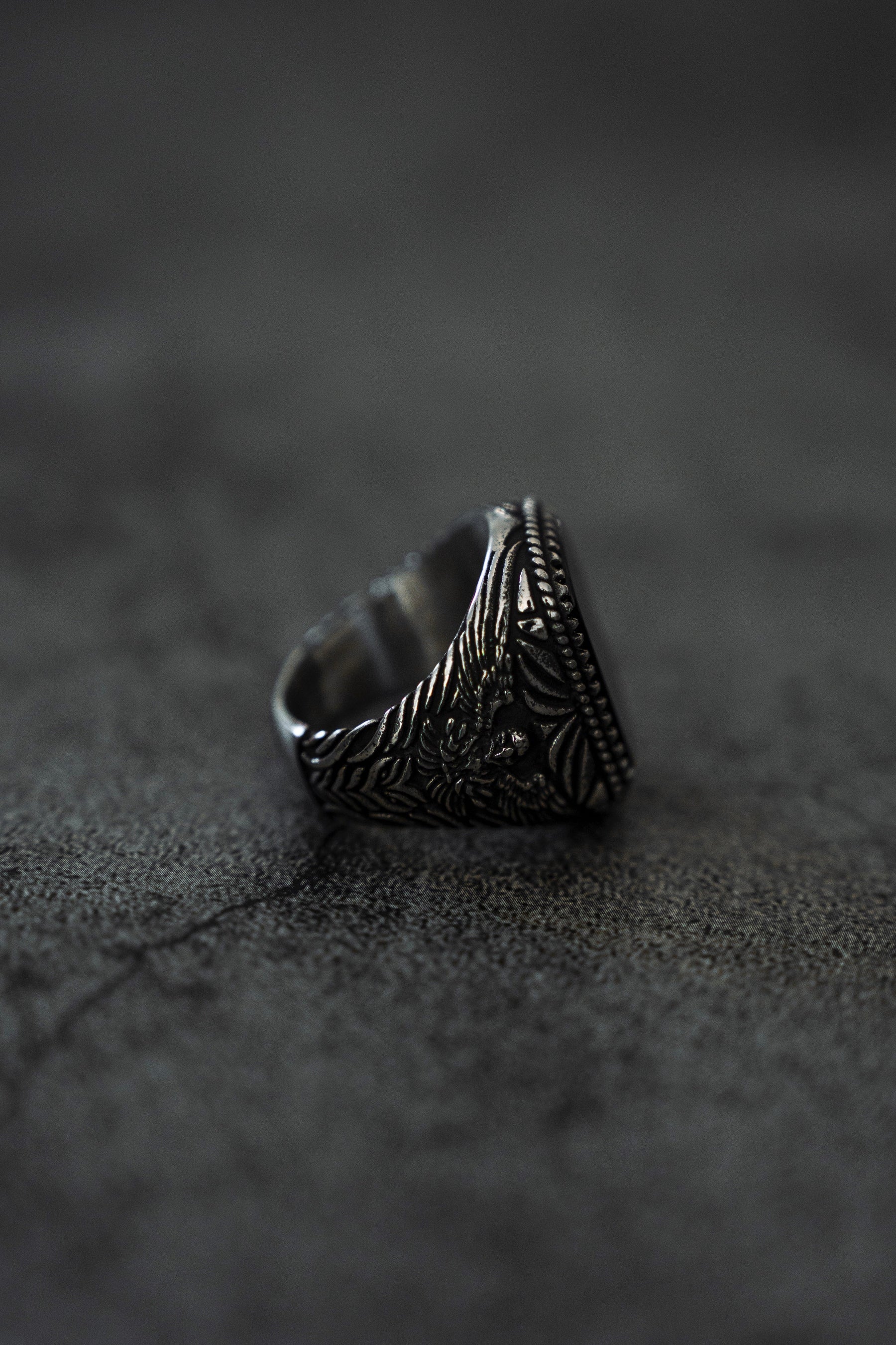 The Monarch Ring by Relentless Betrayal, an ornate stainless steel piece with intricate floral engravings, is displayed on a textured dark surface. Slightly tilted, it highlights its detailed design and embodies a captivating transformation in craftsmanship.