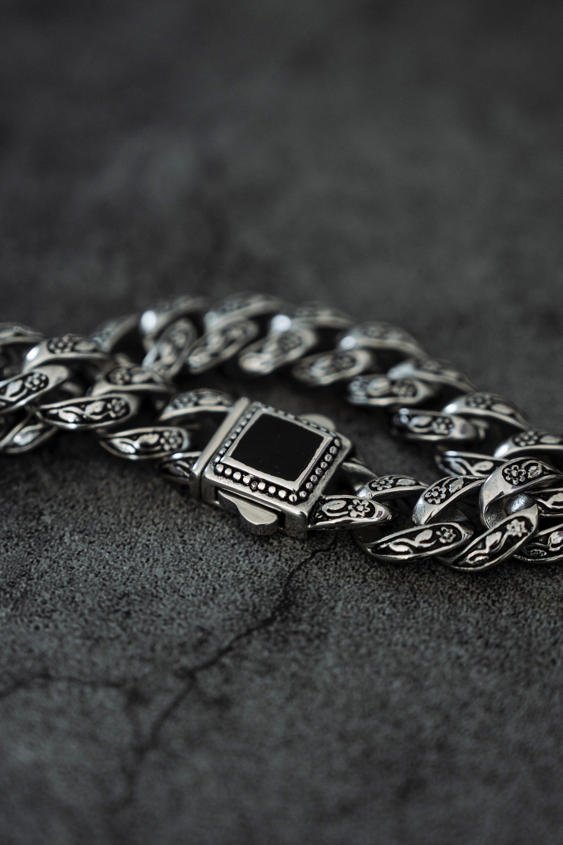 A close-up of The Monarch Bracelet by Relentless Betrayal features an intricately designed stainless steel chain with detailed links and a decorative clasp, set against a textured dark gray background.