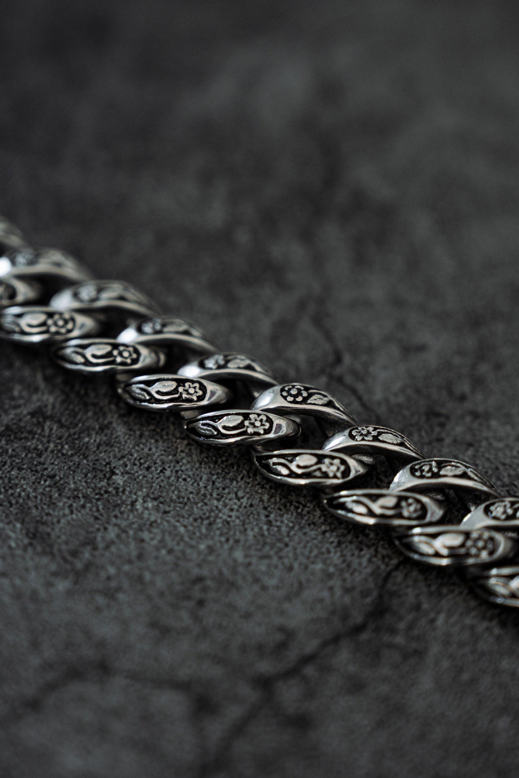 Close-up of The Monarch Bracelet by Relentless Betrayal, crafted in silver-toned stainless steel with intricate floral engravings on each link. Laid on a textured dark gray surface, this bracelet embodies transformation through its detailed pattern.