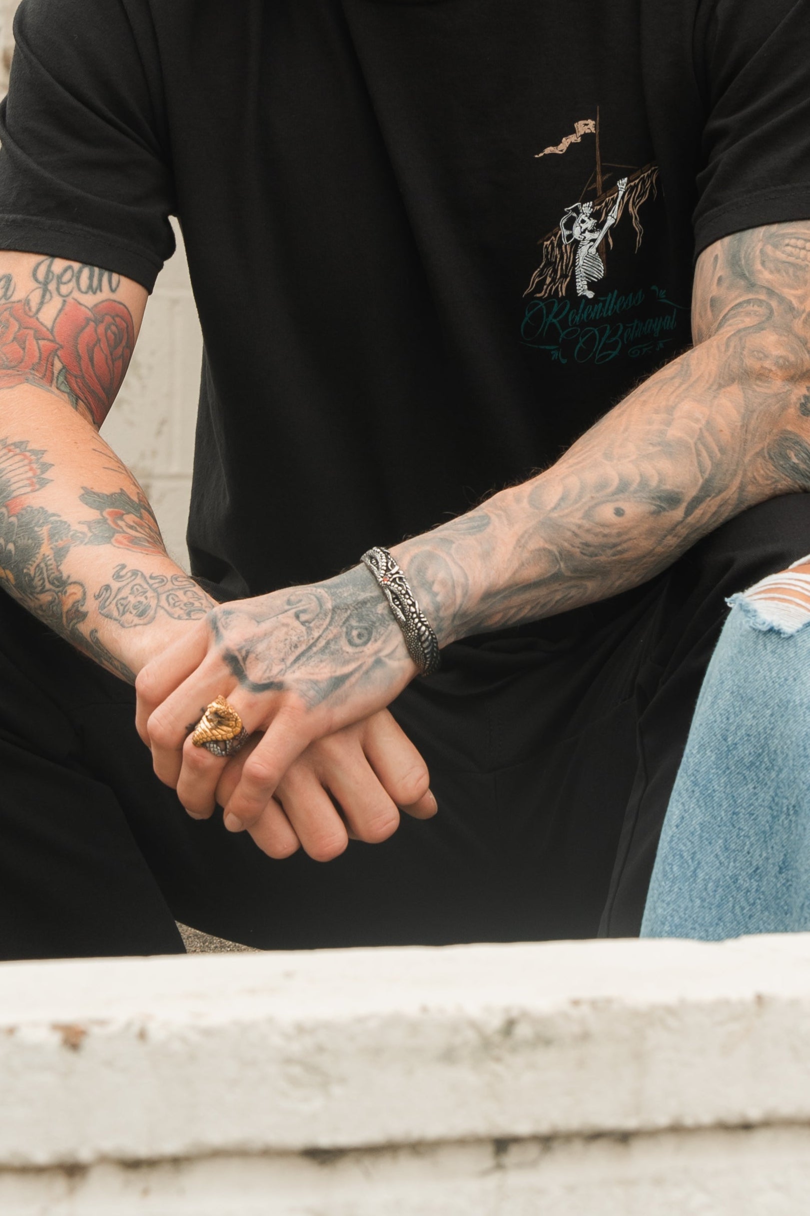 A person with tattooed arms sits wearing a black T-shirt and ripped jeans. Among their bracelets, the Relentless Betrayal Holy Grail Bracelet stands out. A ring adorns their fingers, but their face remains hidden from view.
