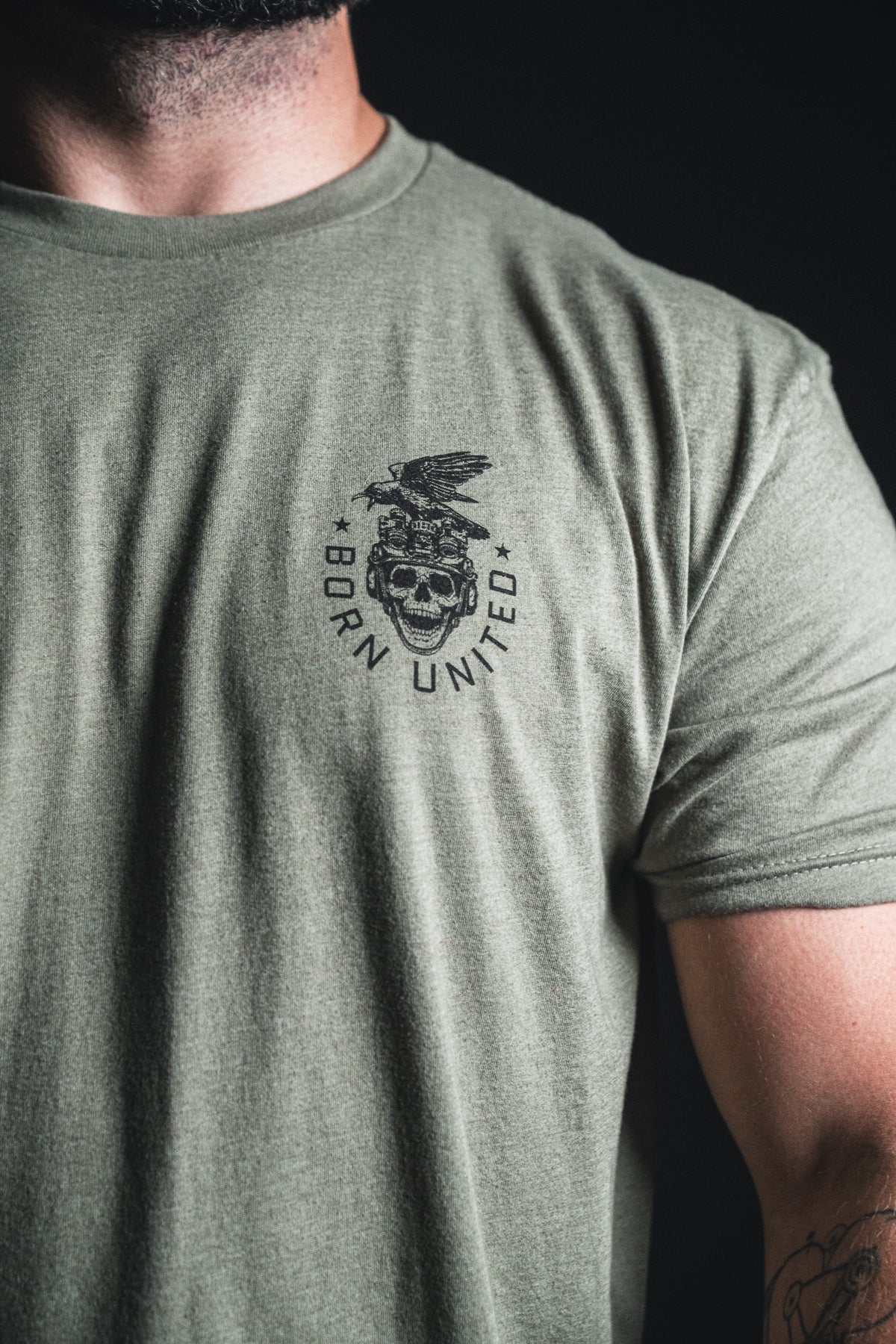 A person wearing an olive green “Only the Dead” t-shirt from Born United Private Label, featuring a skull, eagle, stars, and the words Born United on the left chest—echoing themes of remembrance and history.