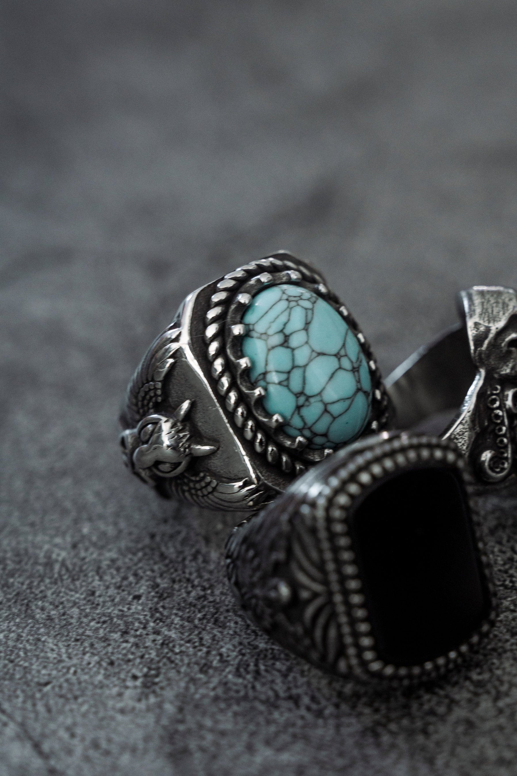 Close-up of three ornate rings on a textured gray surface: an antique heirloom with a turquoise stone and braided silver border; another displays a black stone. The third, by Relentless Betrayal, has intricate detailing like the Royal Guardian Ring but without stones.