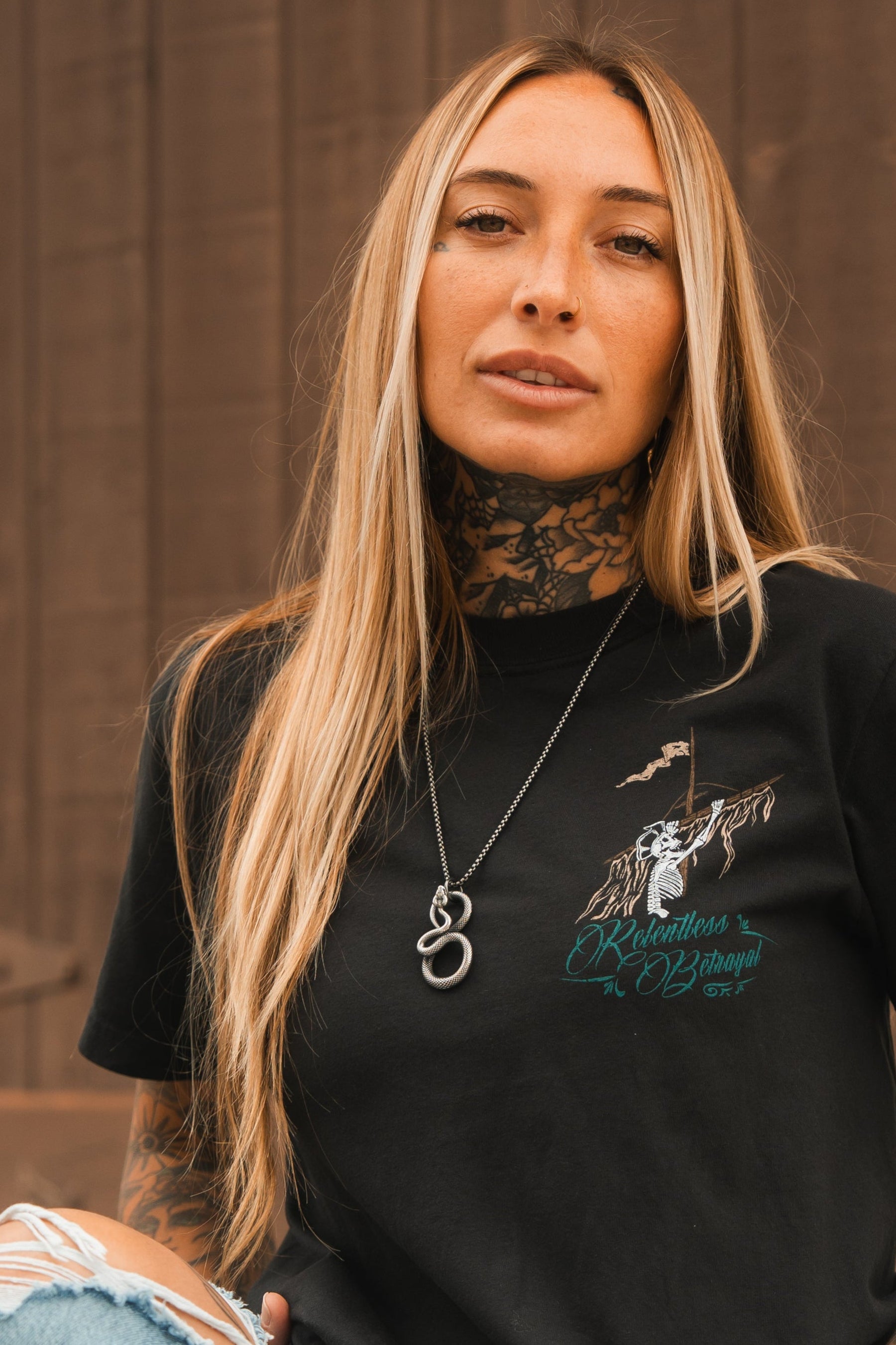 A person with long blonde hair and tattoos on their neck and arms poses in front of a wooden background, wearing a black T-shirt with a design. They make a statement piece out of the Relentless Betrayals Slither Necklace, embodying serpentine elegance.