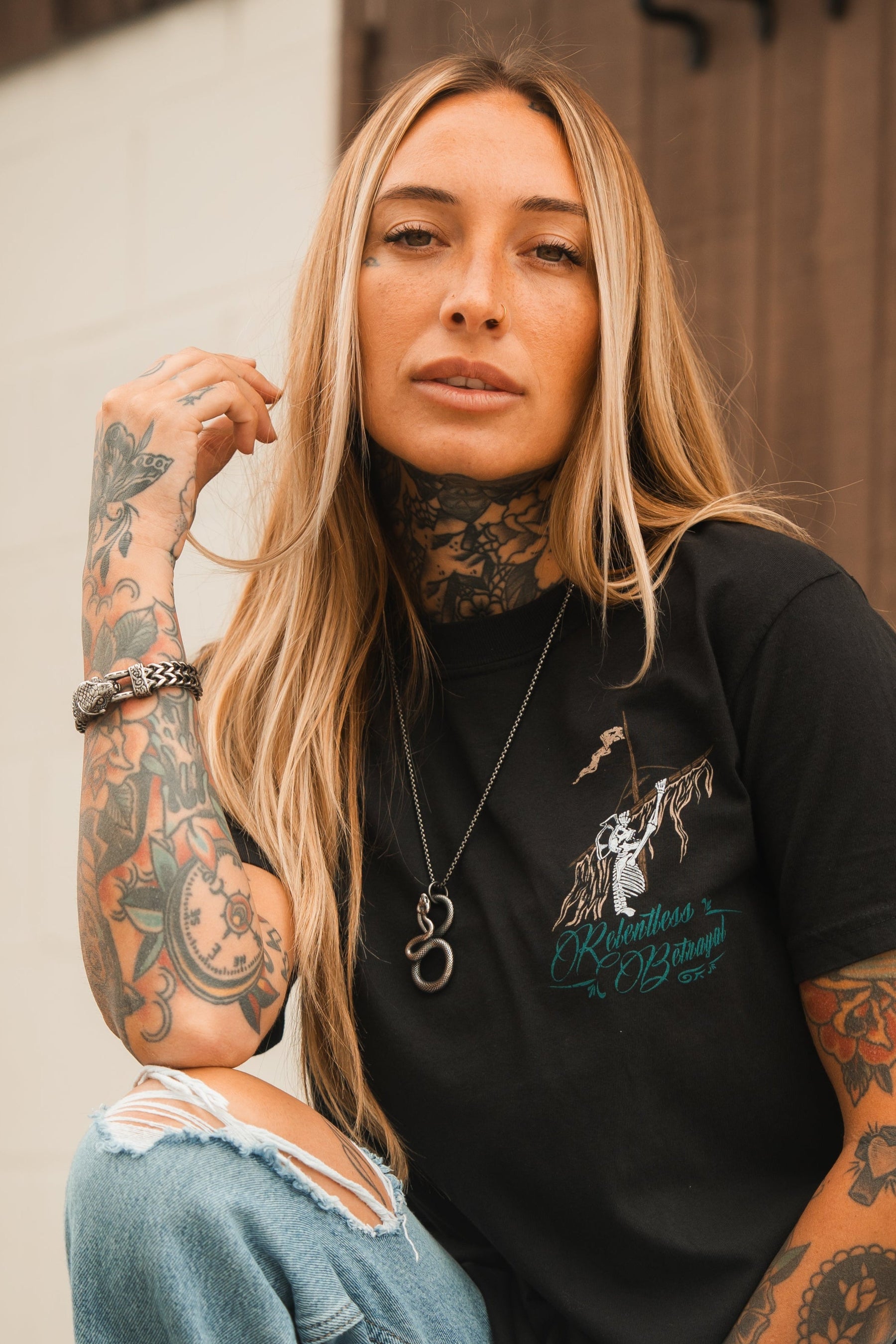 A tattooed woman with long blonde hair sits outside, wearing ripped jeans and a black graphic T-shirt. With a thoughtful expression and her chin resting on her hand, her look is highlighted by a statement piece—the Slither Necklace by Relentless Betrayal—and a watch.