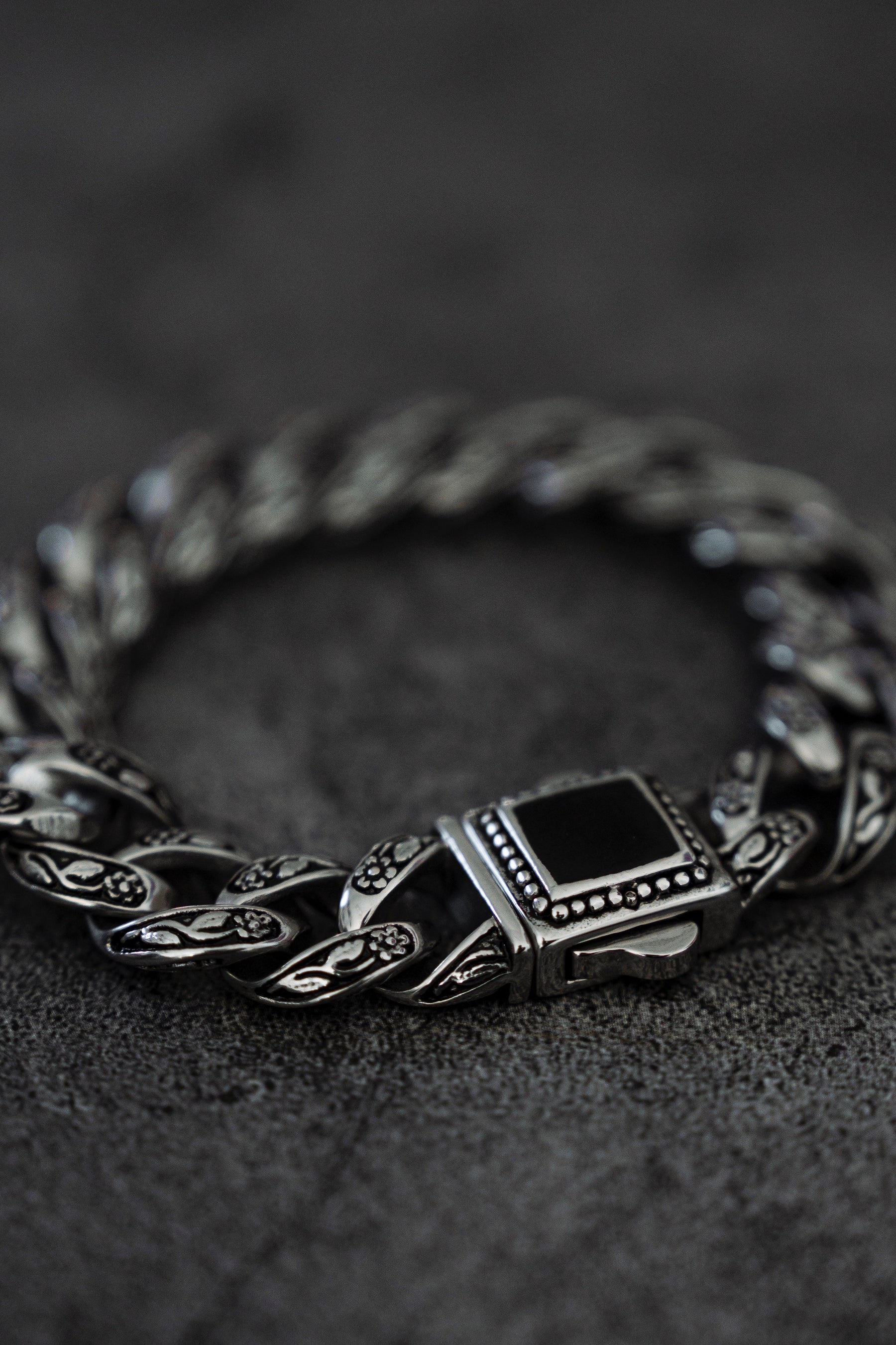 The Monarch Bracelet by Relentless Betrayal features a silver chain made of stainless steel with intricate designs, highlighted by a rectangular black stone centerpiece, elegantly set on a textured dark surface.