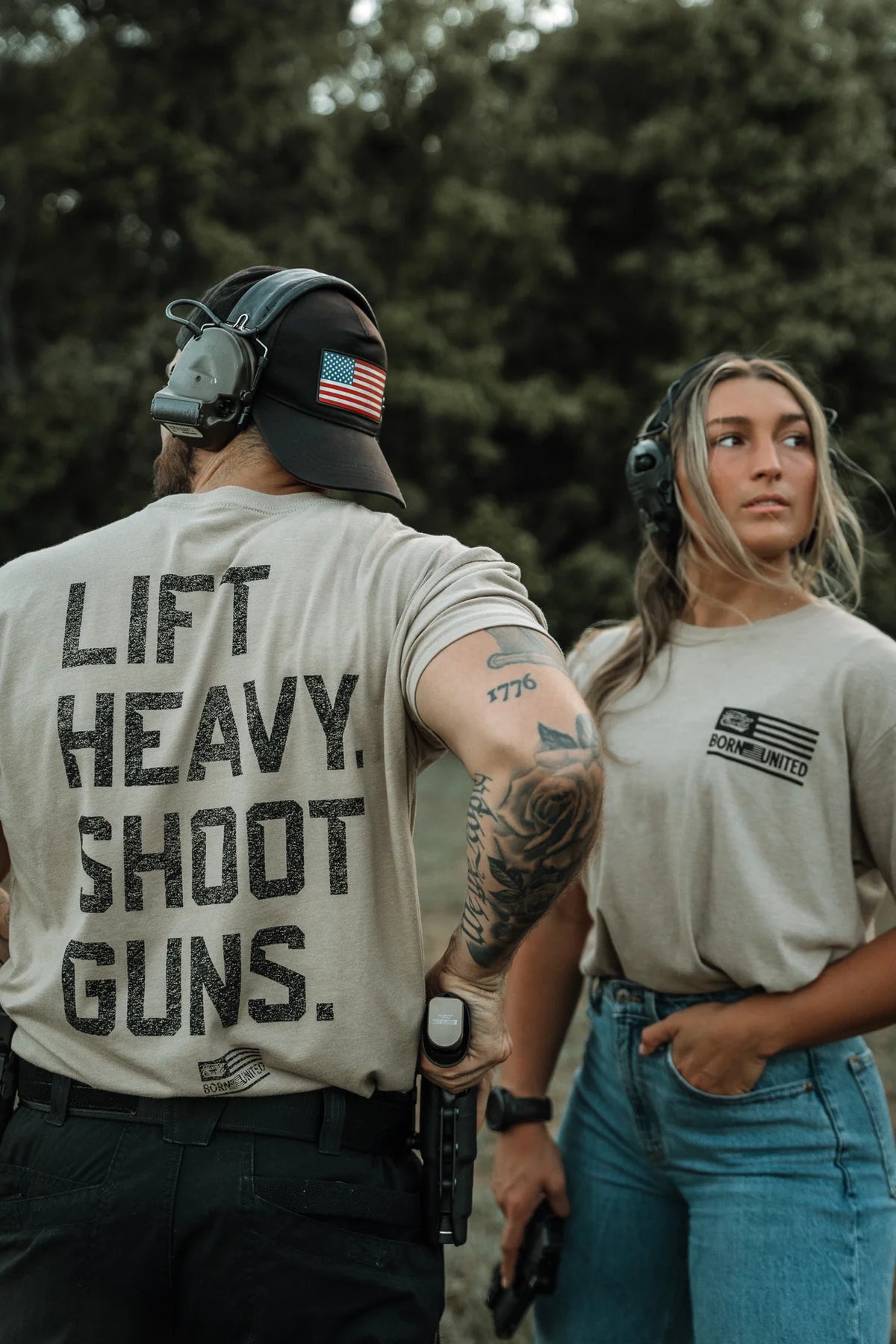 Two people wear Tango Charlie Apparel’s “Lift Heavy. Shoot Guns.- Tan” shirts outdoors. The man, with tattoos, a US flag cap, and a holstered handgun, stands beside a woman holding a pistol and looking to the side.