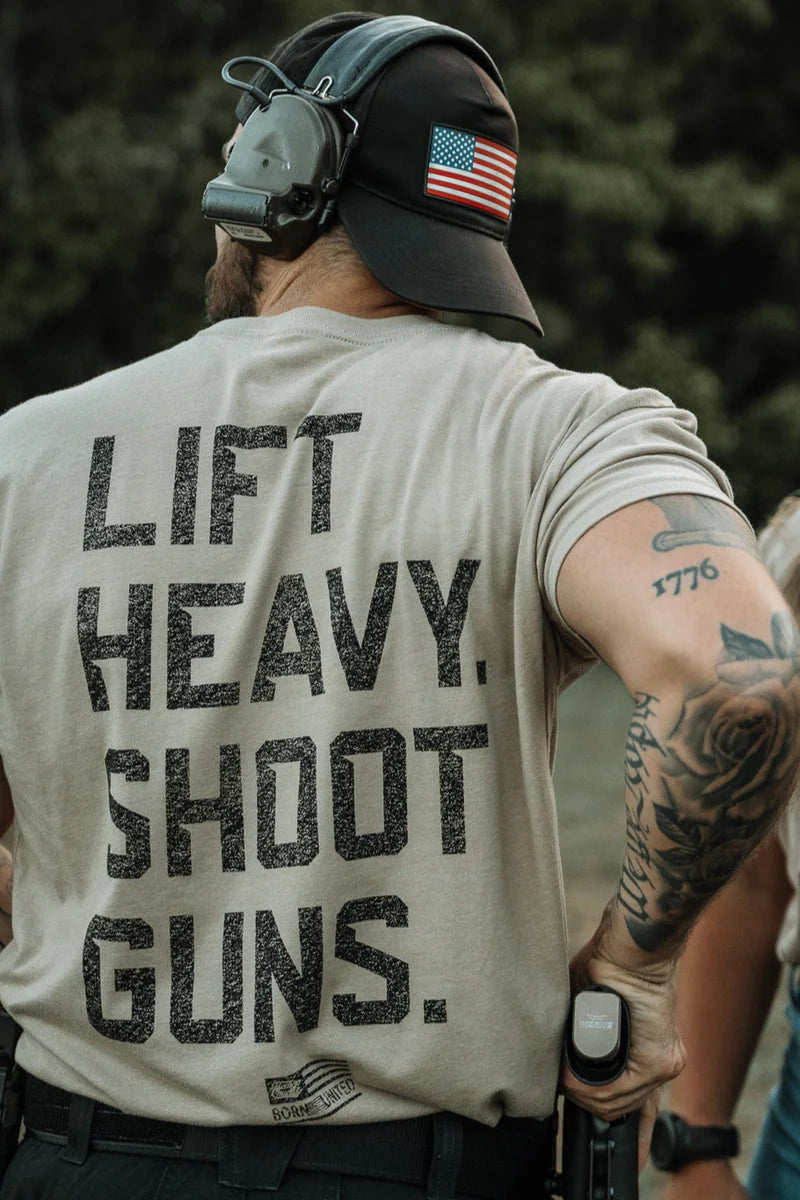 A tattooed person in a headset wears a Tango Charlie Apparel "Lift Heavy. Shoot Guns.- Tan" shirt and a cap with a U.S. flag patch—perfect high-intensity fitness gear—while standing outdoors, viewed from behind.