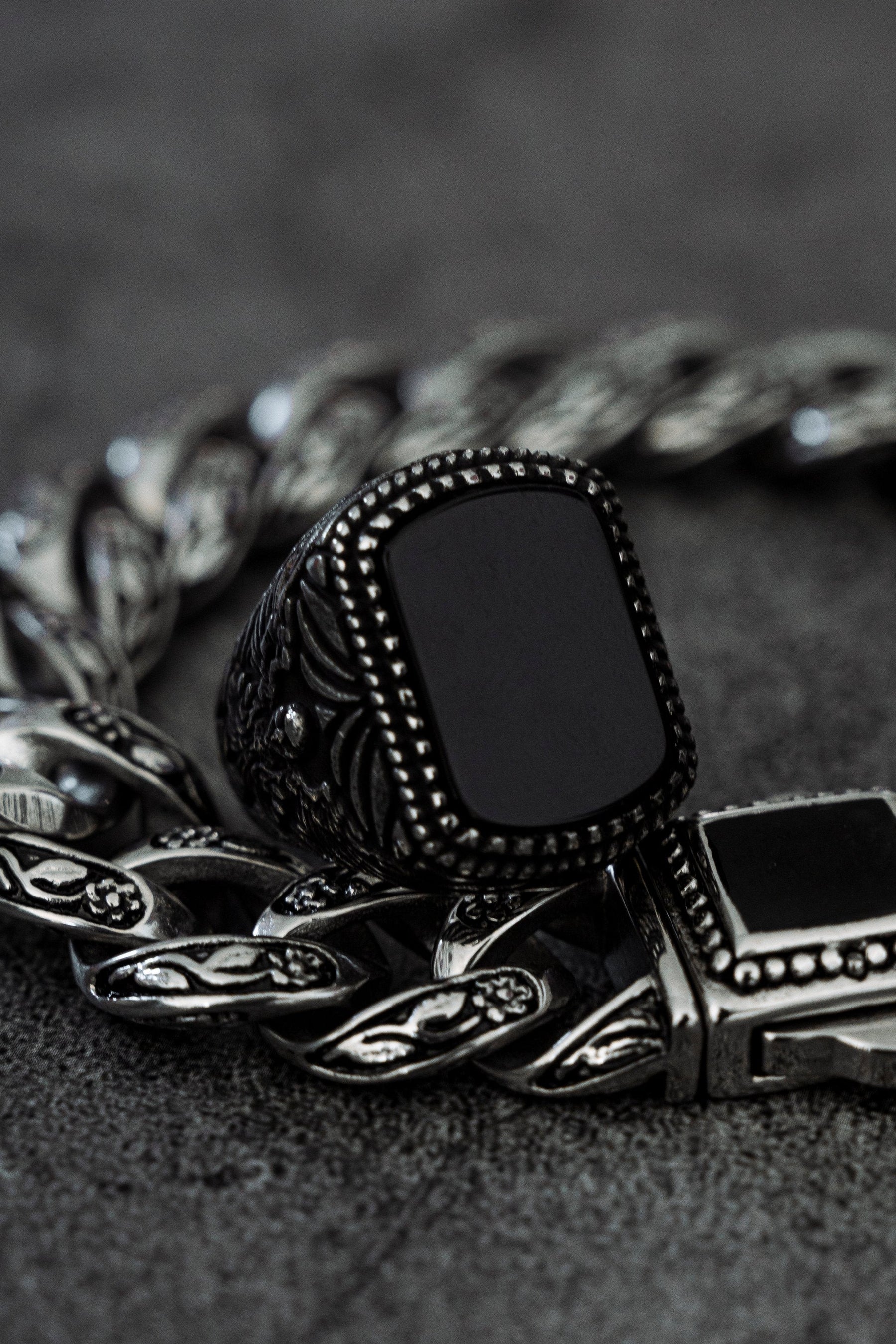 A close-up of Relentless Betrayals stainless steel Monarch Ring, featuring a black rectangular stone, paired with a matching silver chain bracelet on a textured dark gray surface, highlights their intricate patterns and polished finish, embodying elegance and transformation.