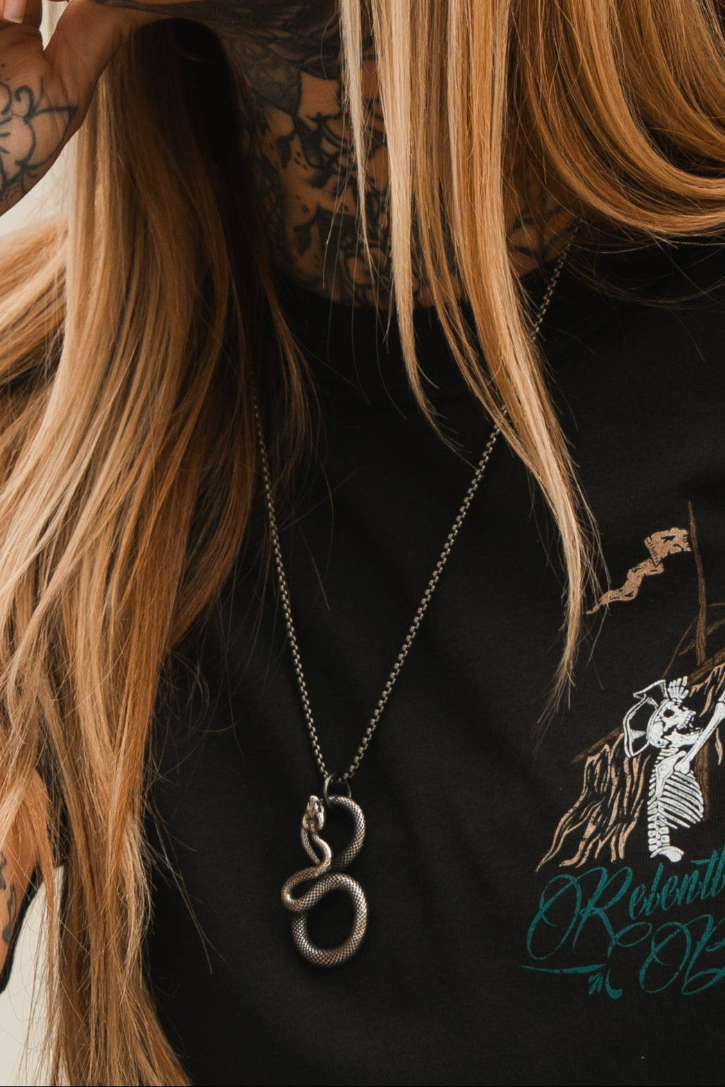 A person with long blonde hair, tattooed arms, and a black shirt with an illustrated design exudes serpentine elegance. The Slither Necklace by Relentless Betrayal graces their neckline as a true statement piece.
