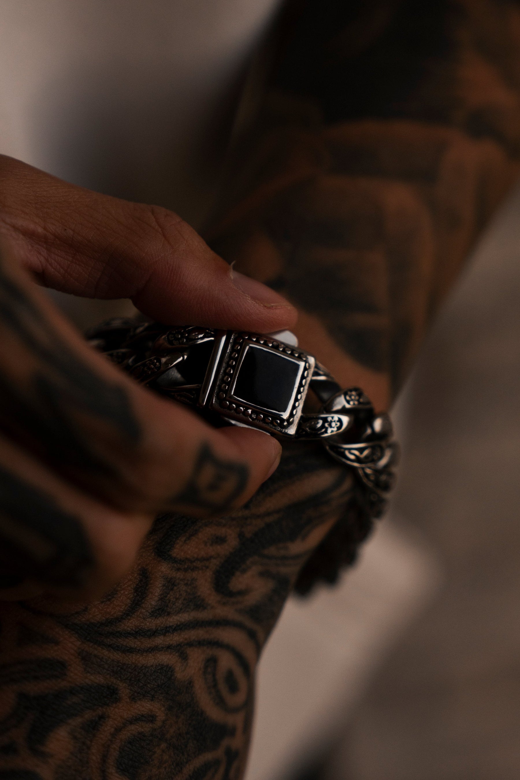 In dim light, tattooed hands carefully fasten Relentless Betrayals Monarch Bracelet, made of stainless steel with a square black stone, around their wrist—a simple adornment that transforms style and elegance.