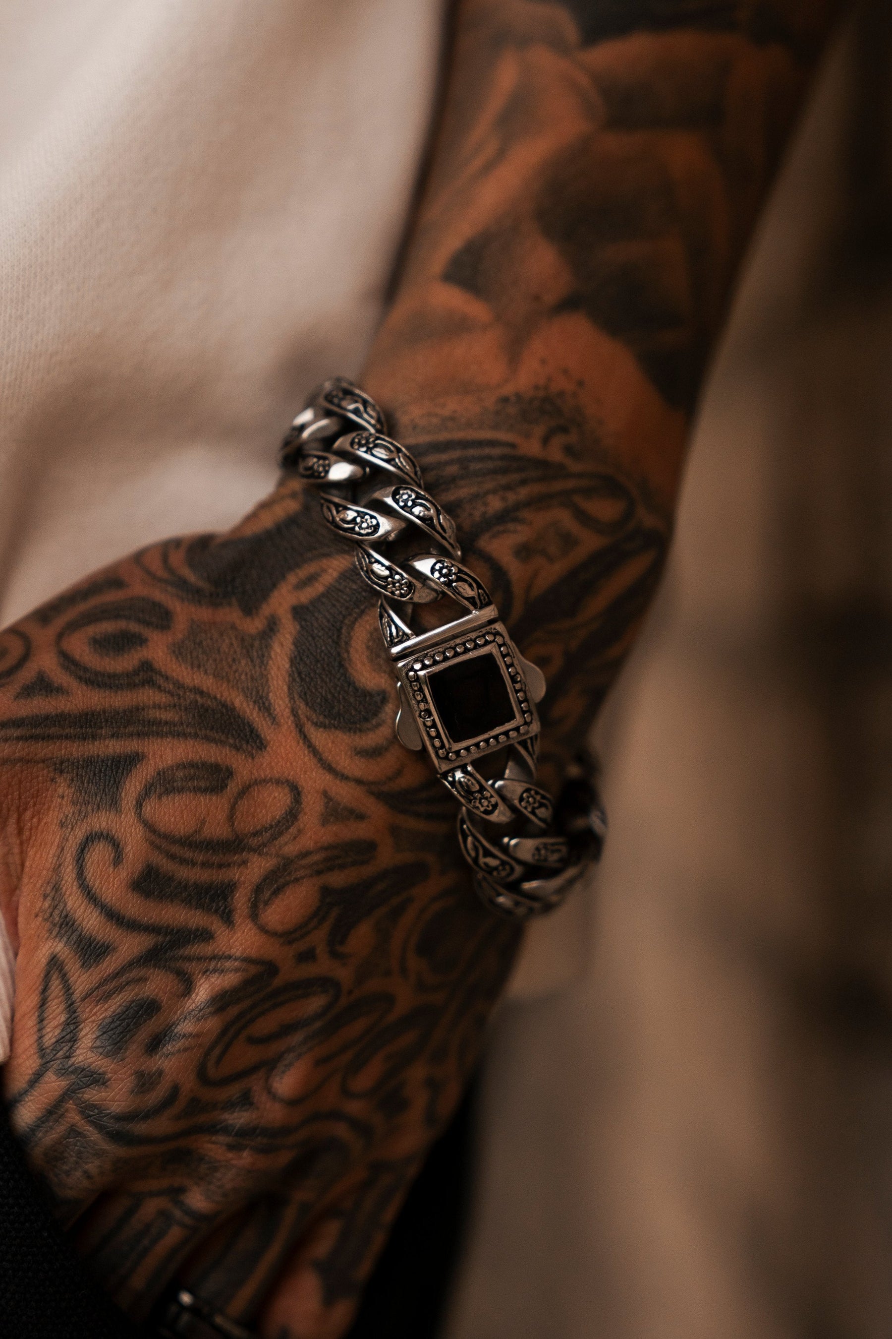 A close-up of a tattooed arm showcases Relentless Betrayals Monarch Bracelet, featuring ornate stainless steel and a central square dark stone. Intricate tattoos flow across the forearm and hand, symbolizing transformation.