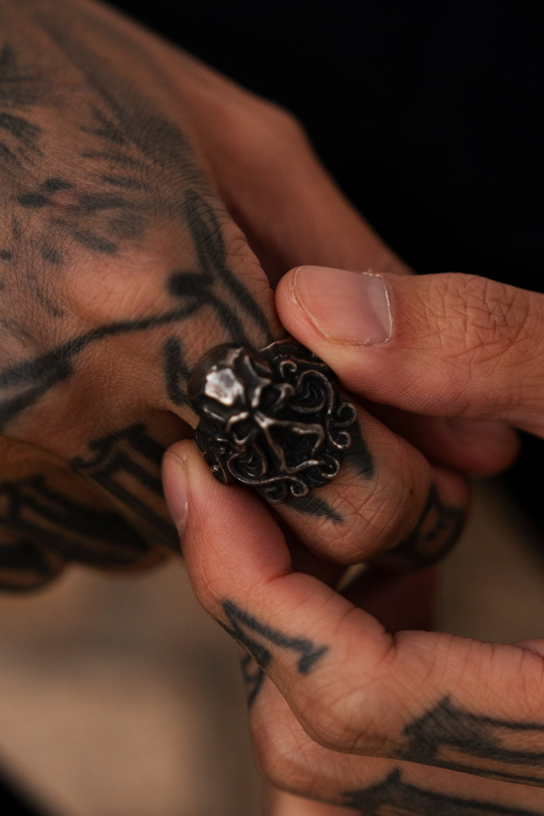 A close-up features a finger with intricate black tattoos, adorned by The Kraken Ring from Relentless Betrayal—an elaborate handcrafted silver skull design. A blurred background highlights the hand and the rings Kraken-inspired details.