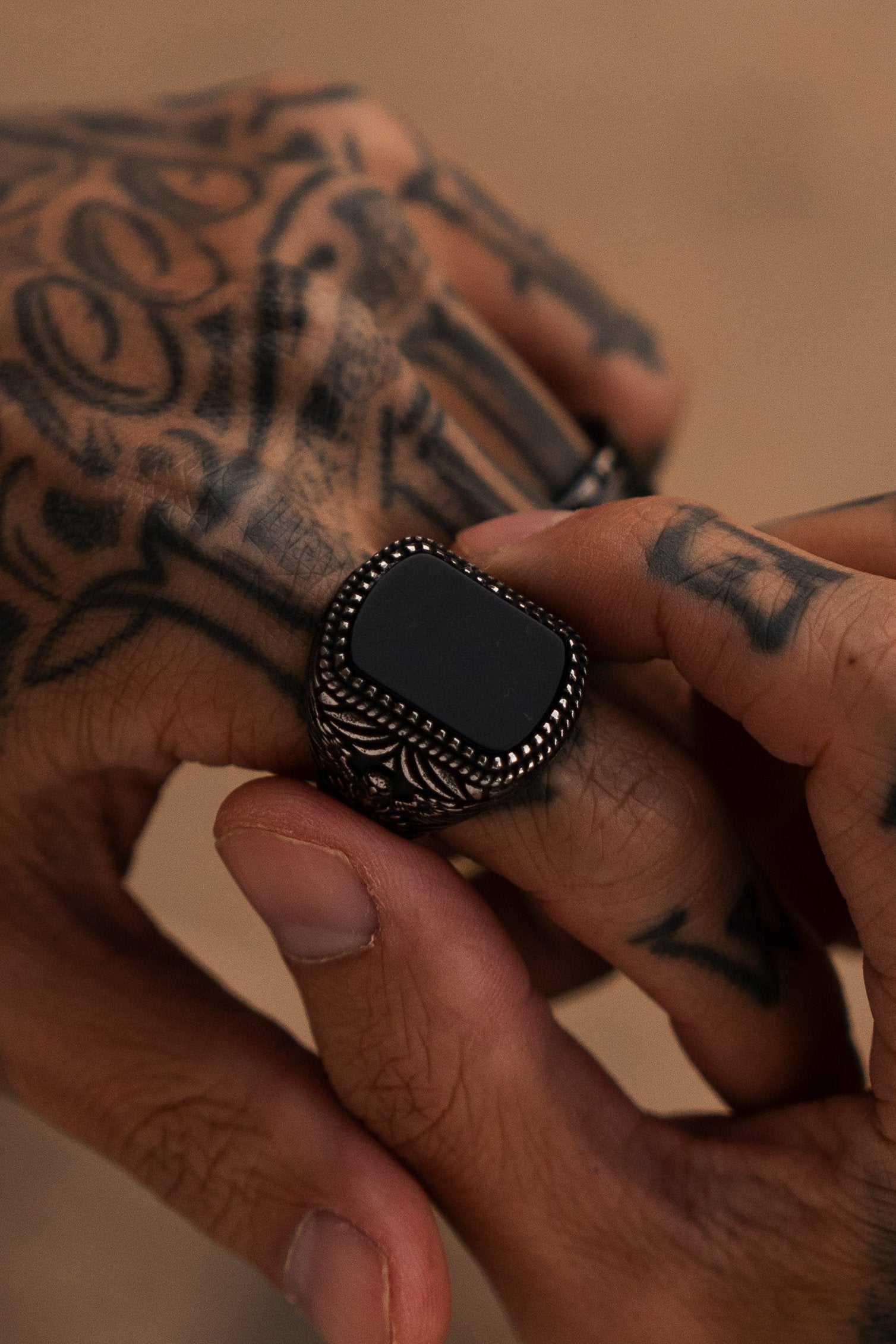 Close-up of tattooed hands adjusting The Monarch Ring by Relentless Betrayal, showcasing a prominent black stone in intricate stainless steel. Tattoos with patterns and symbols suggest transformation against a softly blurred background.