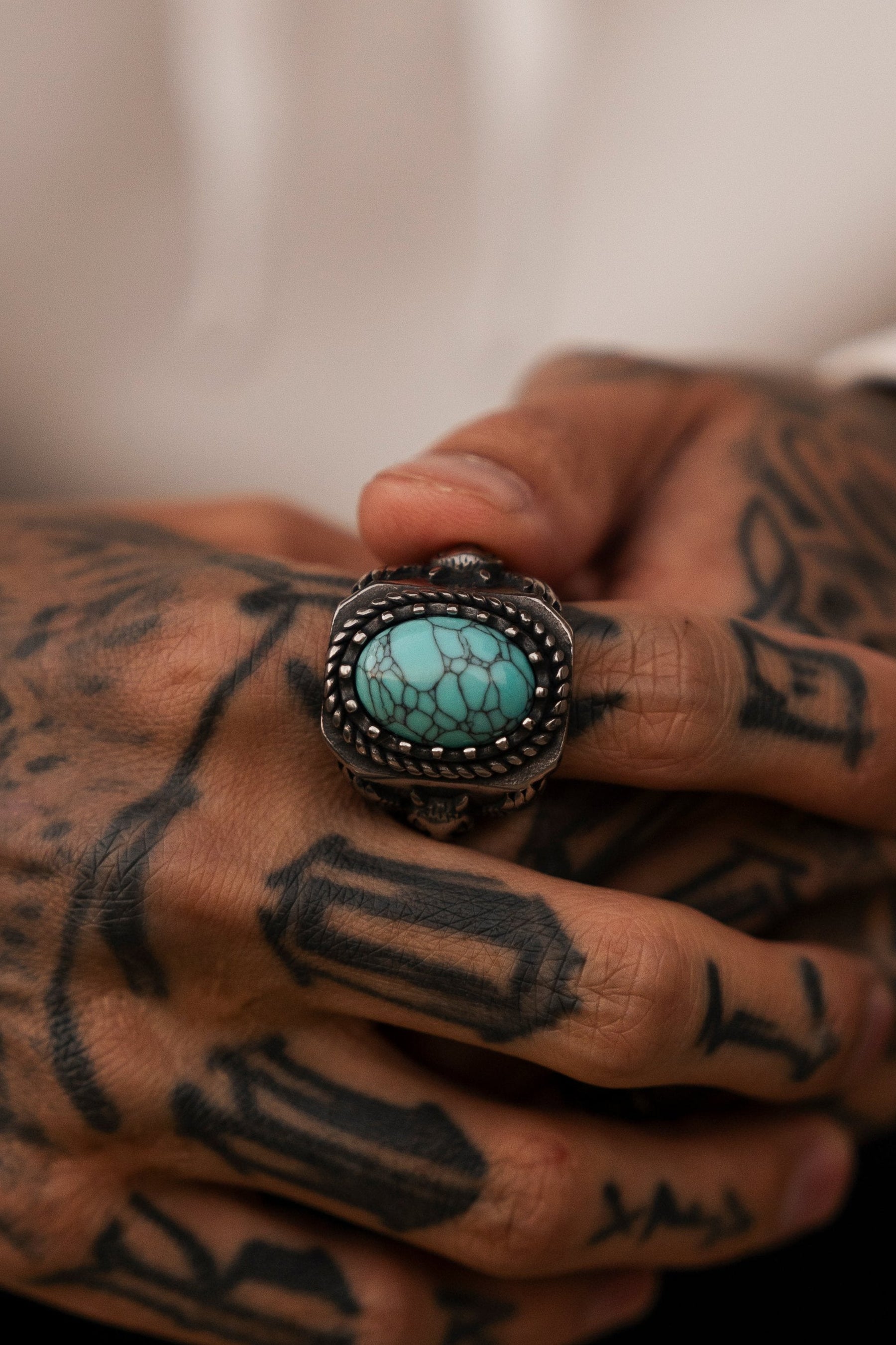 Tattooed hands display the ornate, handcrafted stainless-steel Royal Guardian Ring by Relentless Betrayal, featuring an oval turquoise stone. The tattoos consist of geometric shapes and patterns. A blurred background shifts focus to the hands and ring.