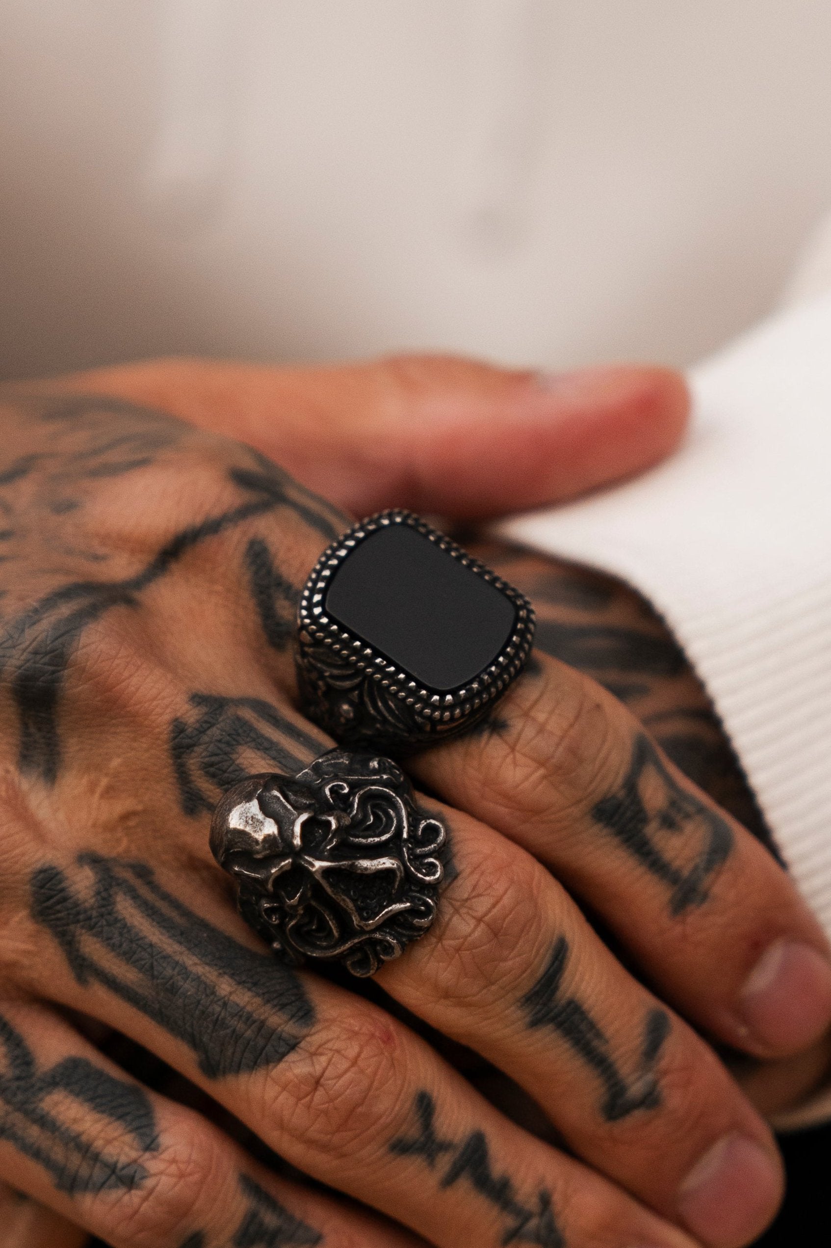 A tattooed hand, adorned with two bold rings, stands out against a white sleeve. The Monarch Ring by Relentless Betrayal features a black stone with an ornate border, while the other ring displays an intricate design. Both are crafted from stainless steel, exuding elegance and style.