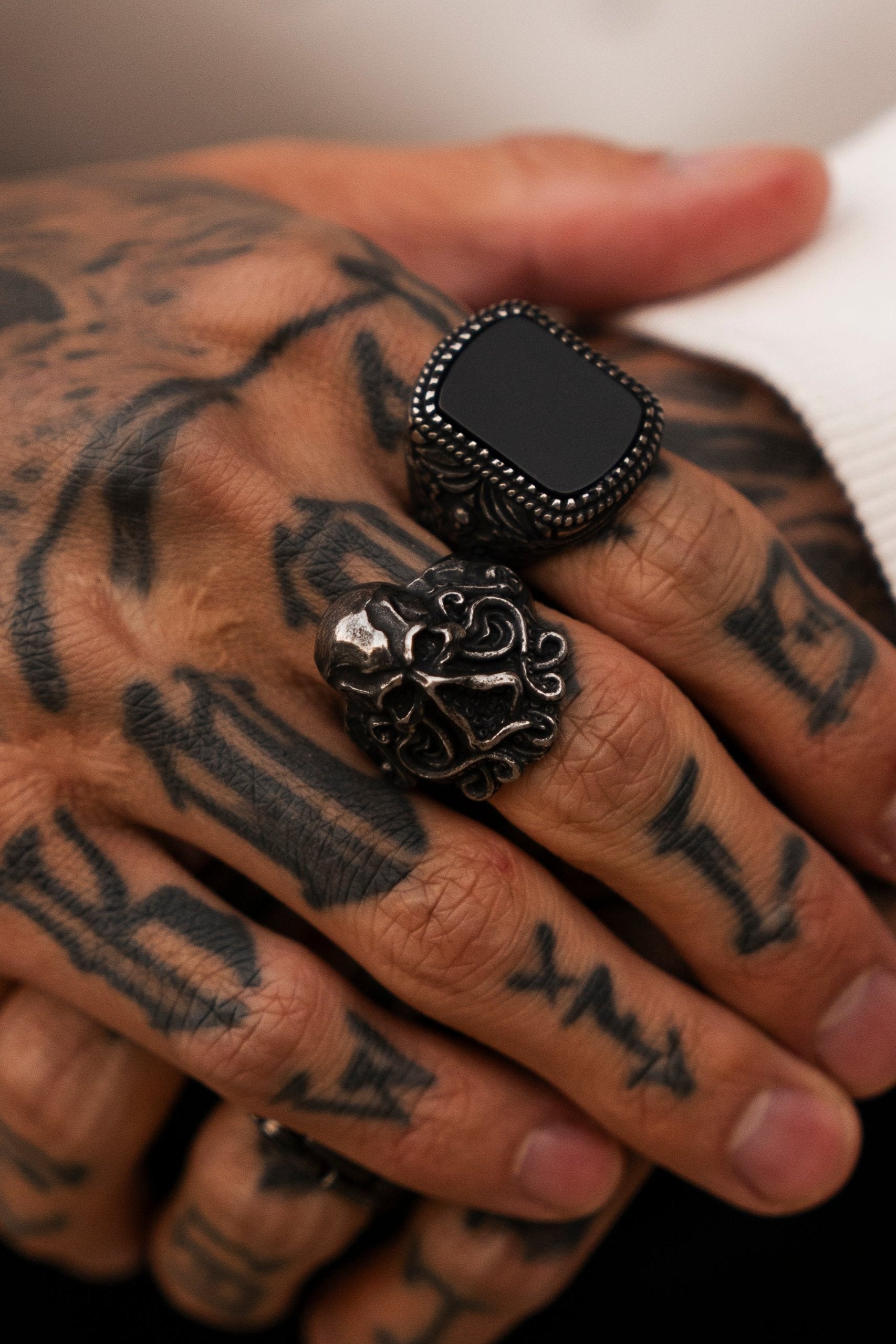 A tattooed hand wearing Relentless Betrayals ornate handcrafted rings: a dark square stone, a detailed lions face, and The Kraken Ring in gleaming stainless steel. The mystique of The Kraken Ring shines near white fabric.