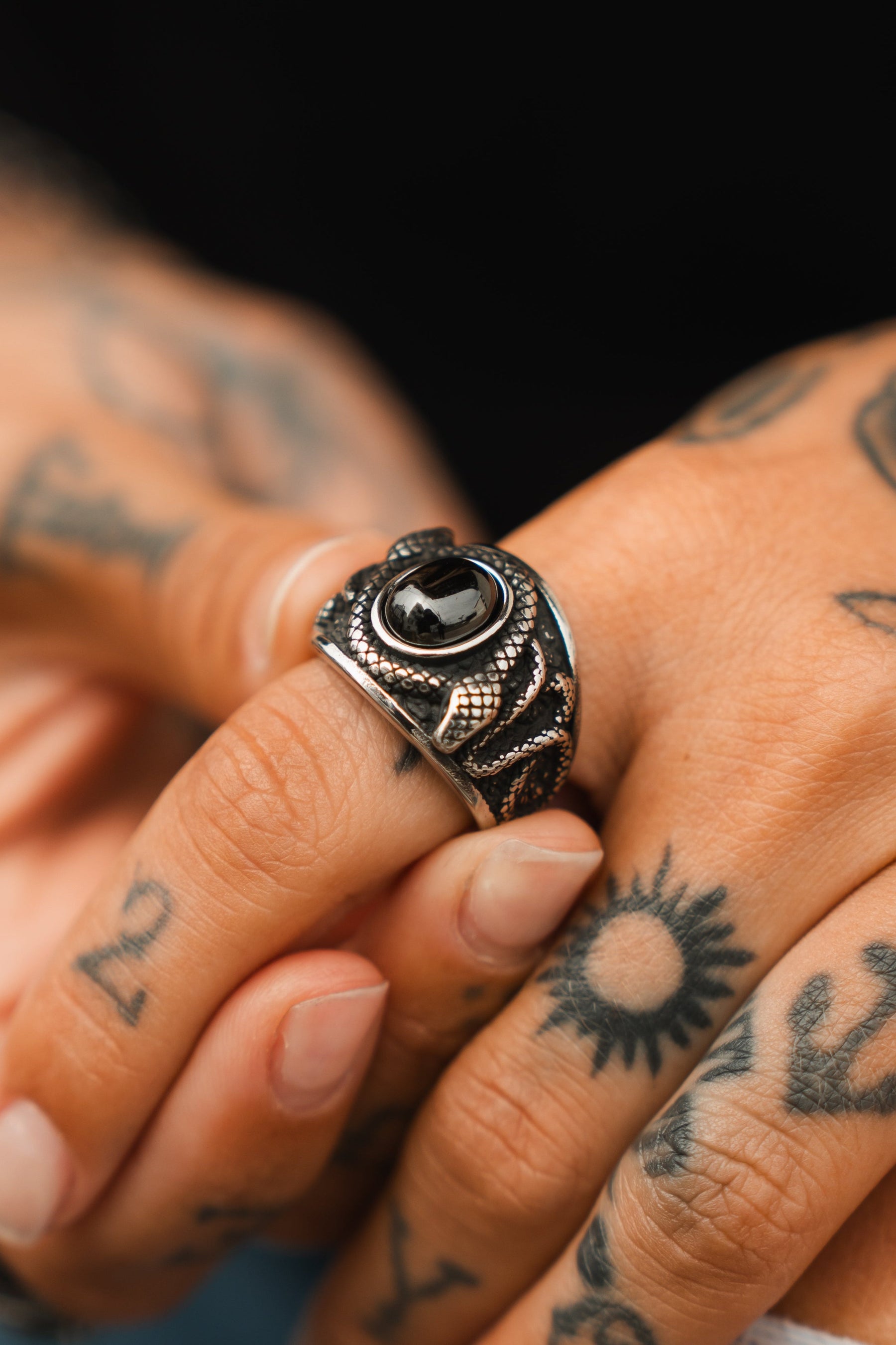 A person dons Relentless Betrayals Onyx Rebirth Ring, a silver handcrafted piece with a black stone and intricate engravings. Set against a dark backdrop, the stainless steel ring complements their tattooed fingers, featuring sun shapes and numbers.