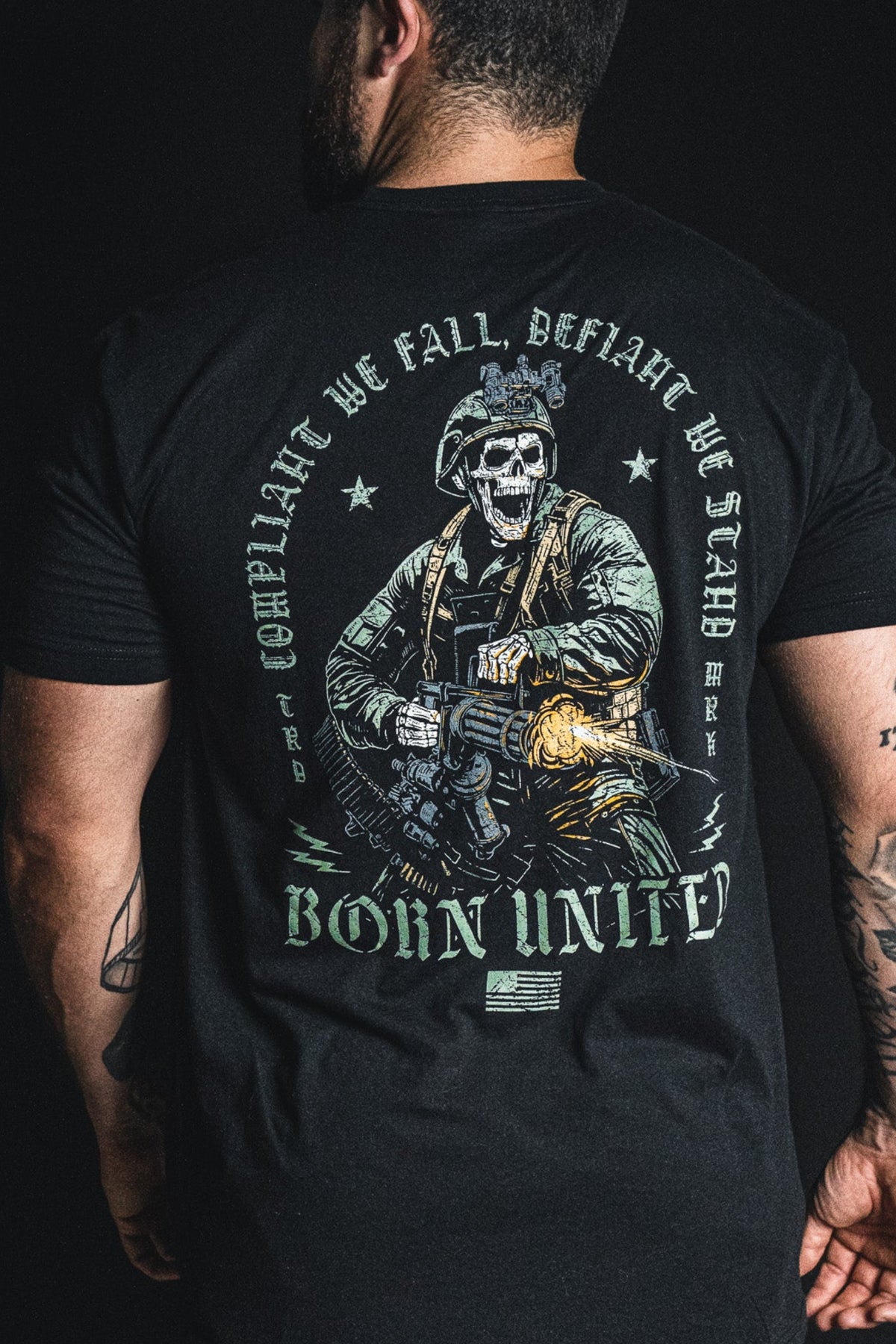 A man viewed from behind wearing a black graphic t-shirt. The back of the shirt features a large distressed print of a skeleton soldier holding a rifle, with the text 'COMPliance WE FALL BEFORe WE STAND' and 'BORN UNITE' in distressed green and gray lettering.
