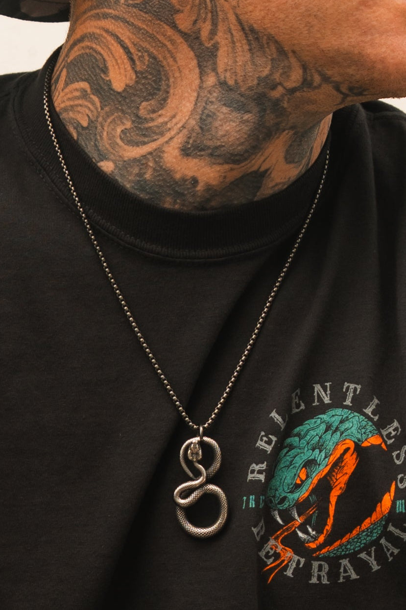 A person wearing a black Relentless Betrayal shirt featuring a snake graphic and the words Relentless Betrayal exudes serpentine elegance with a striking snake tattoo on their neck, making a bold statement with the Slither Necklace.