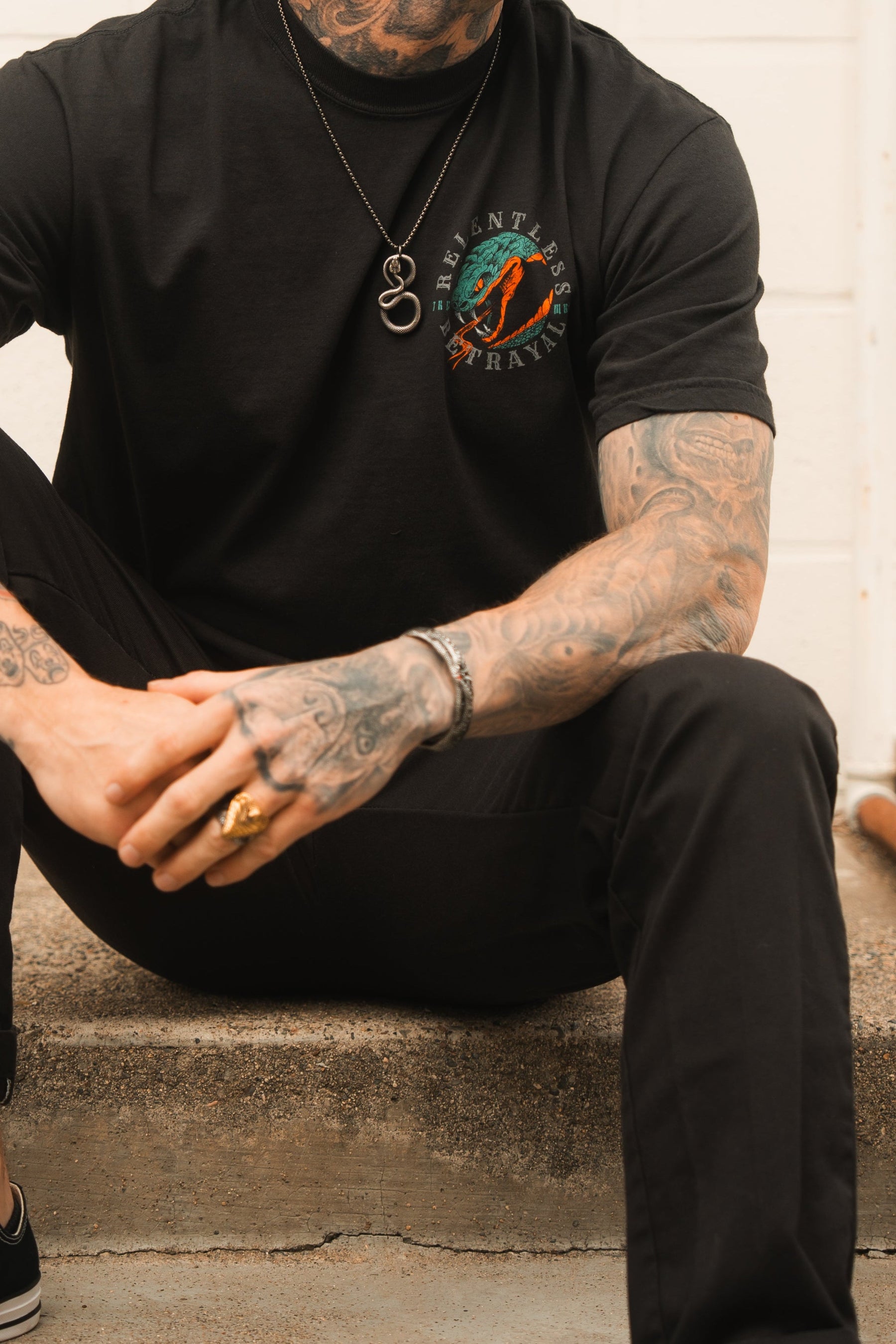 A tattooed person sits on concrete steps, wearing black pants and a t-shirt with a colorful design. Adorned with Relentless Betrayals Holy Grail bracelet and various rings, their hands are clasped. A necklace featuring a ring pendant drapes over the shirt.