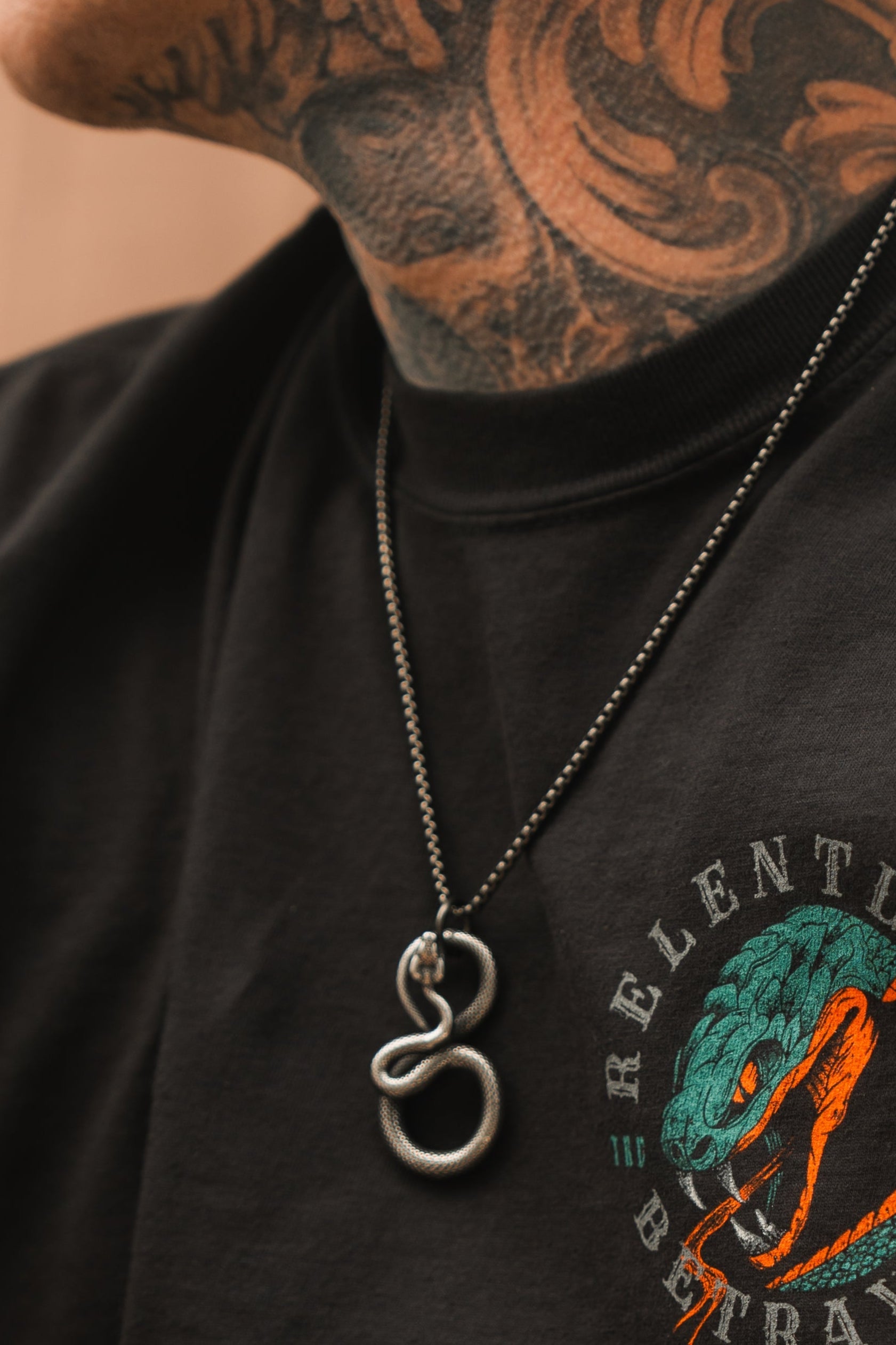 Close-up of a person wearing a black T-shirt featuring a snake graphic with RELENT partially visible. A Slither Necklace from Relentless Betrayal graces their neck, adding serpentine elegance to the detailed tattoos and making a bold statement.